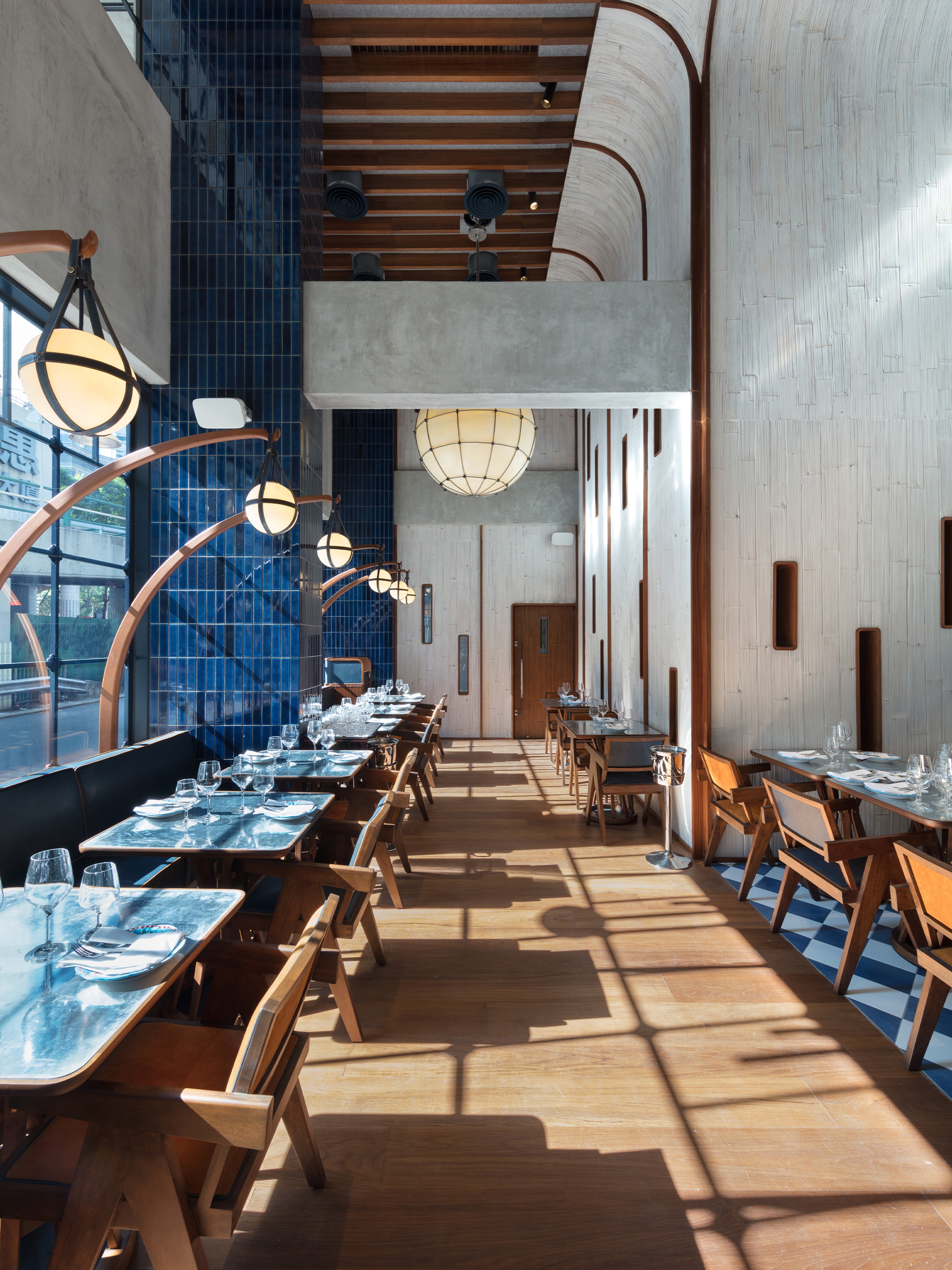Modern restaurant interior with high ceilings, set tables, large street-facing windows, spherical pendant lights, and blue tiled accents with wooden arches.