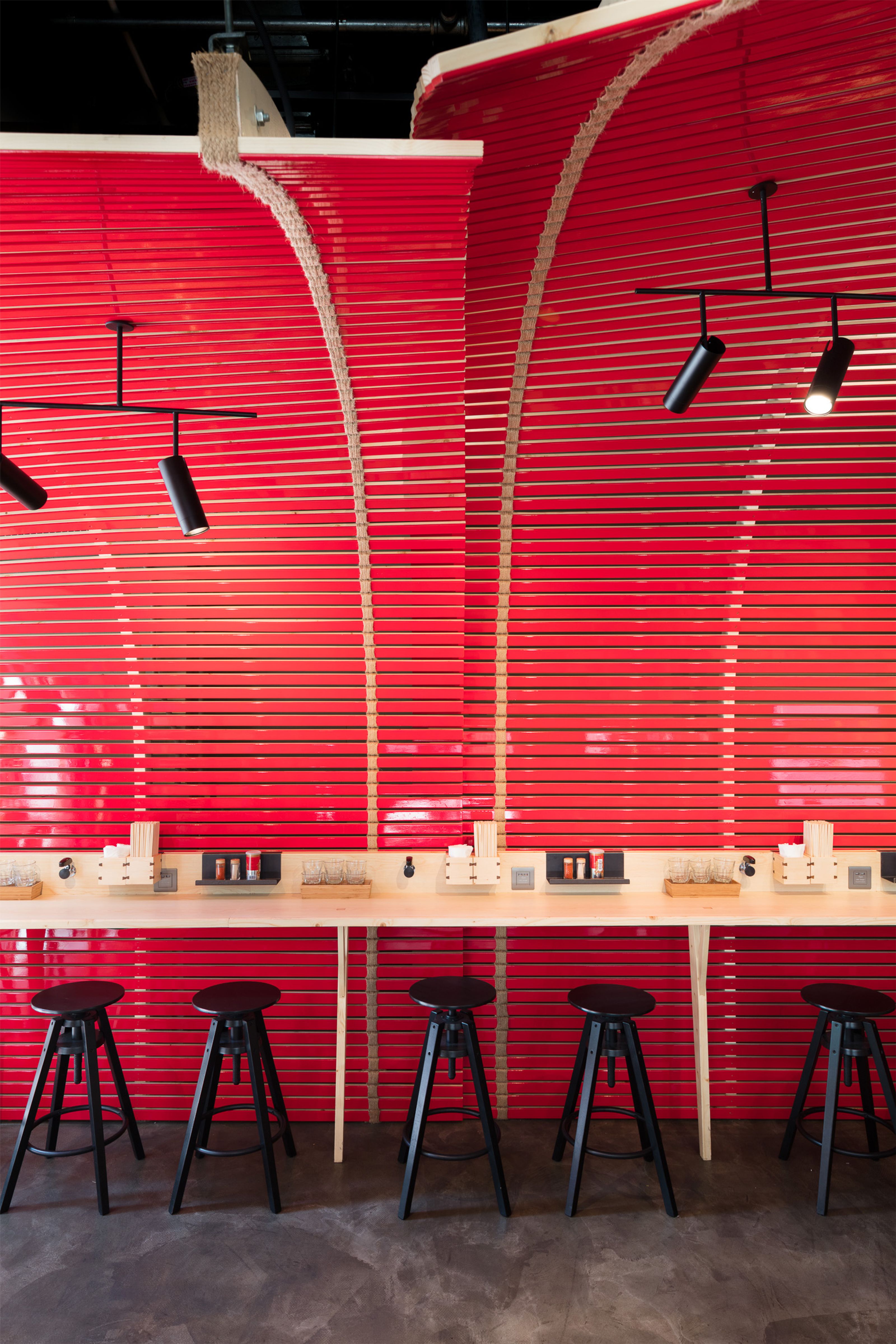 A modern dining area with a vibrant red slatted wall featuring curved natural fiber accents, a light wooden counter, and five black stools.