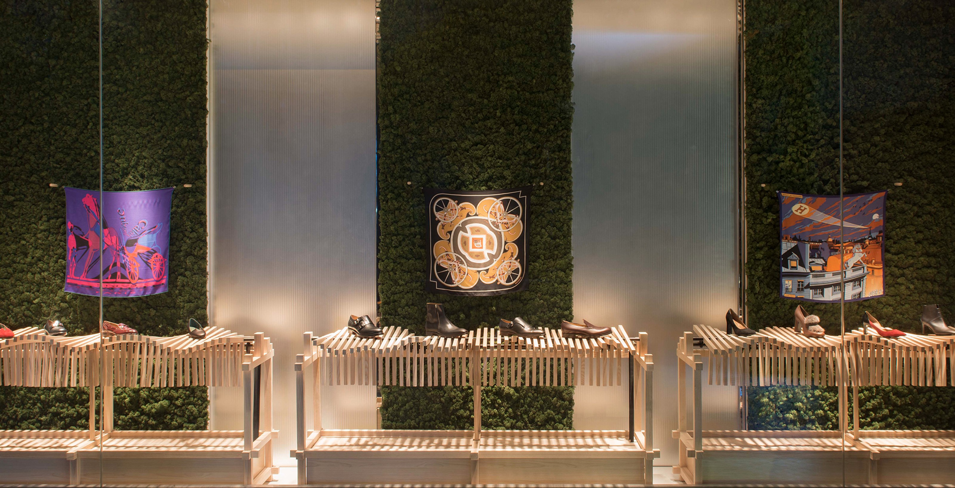 Three colorful silk scarves and various shoes displayed on slatted wooden stands in front of green moss walls in a store window.