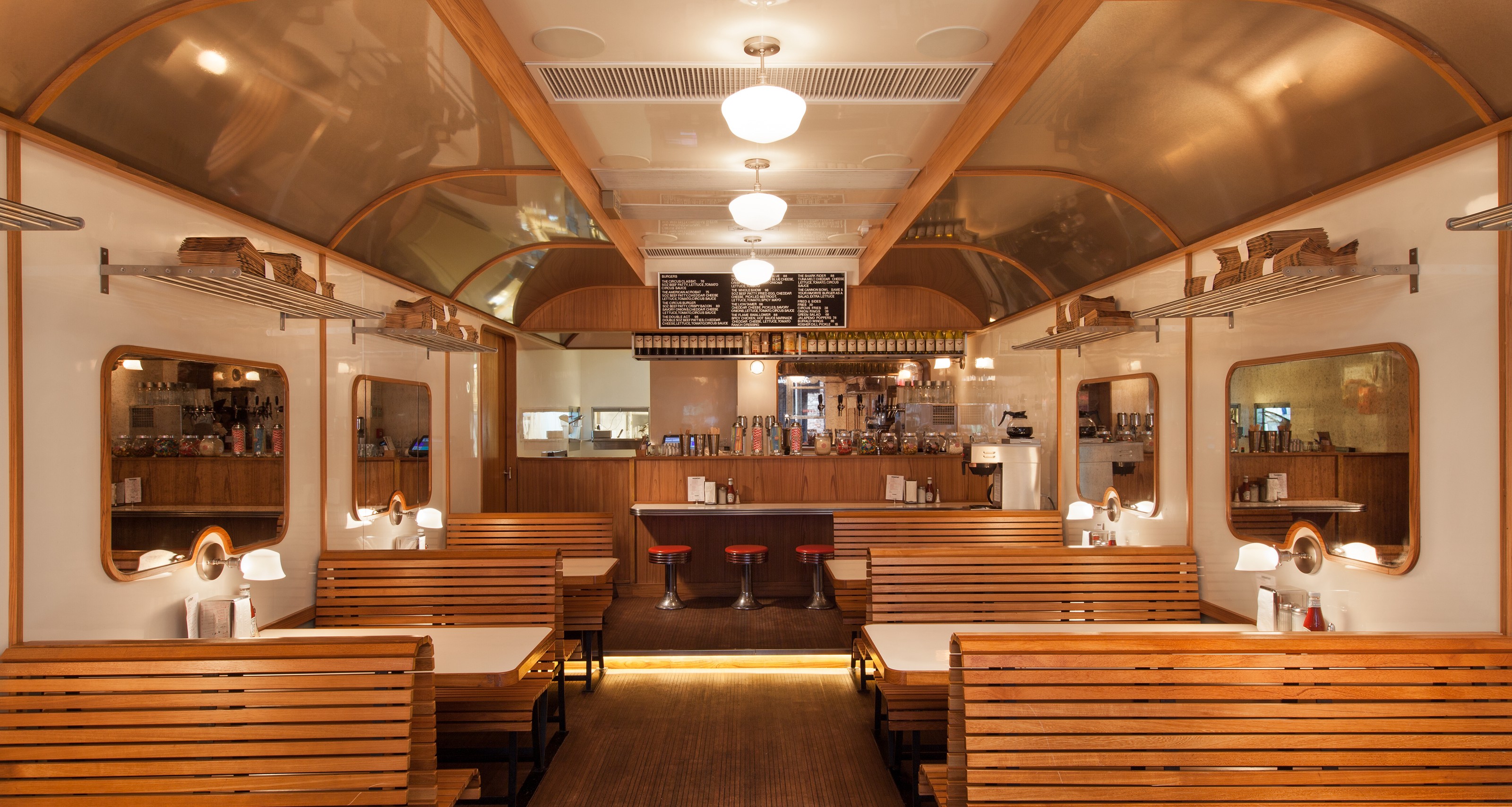 Interior of a train car-themed diner with wooden booths, white tables, overhead racks, and a bar.