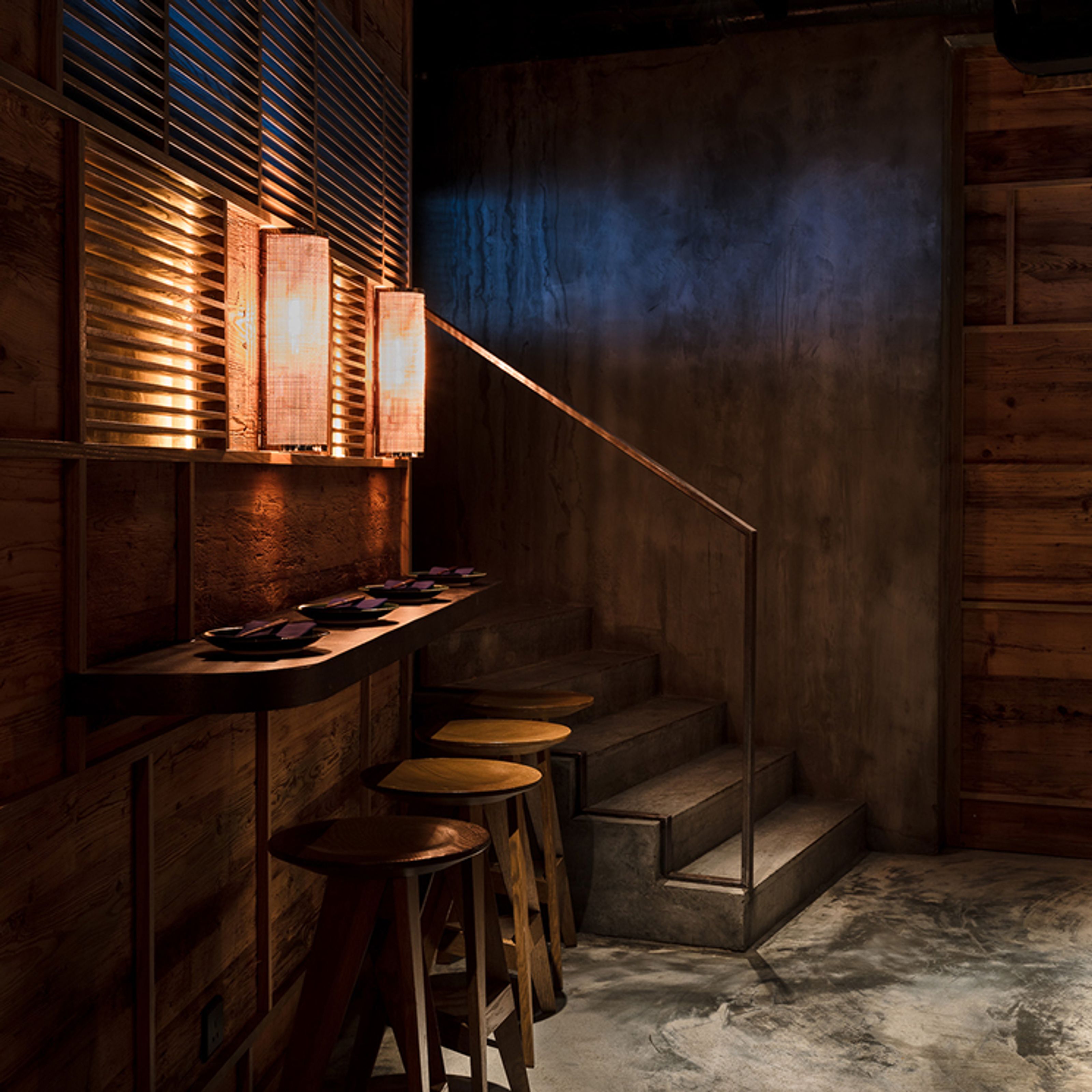 A dimly lit interior with a wooden counter, four stools, three warm vertical lamps, and concrete stairs.