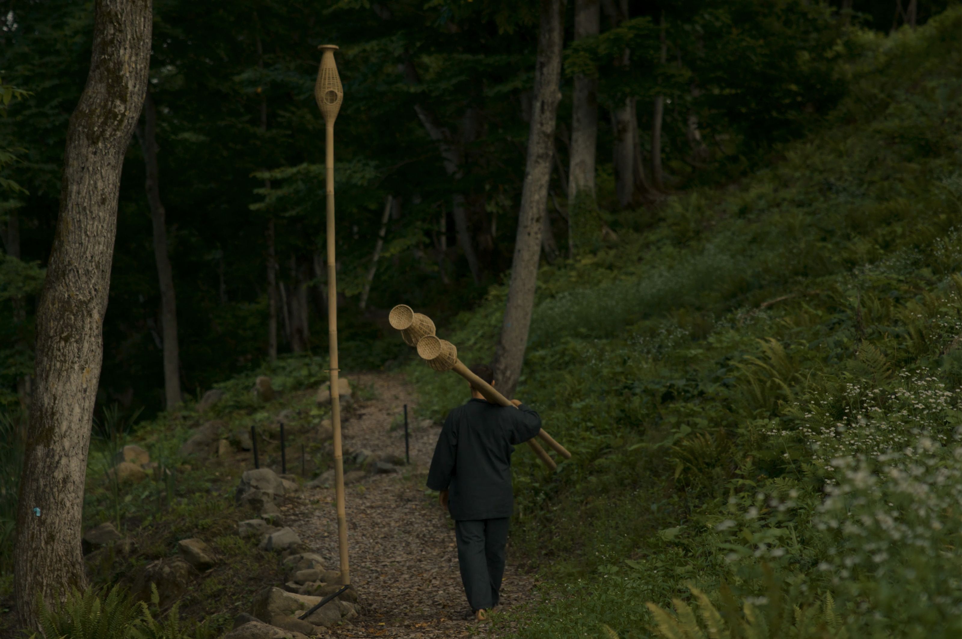 A person walks on a forest path carrying bamboo objects, passing a tall bamboo sculpture.