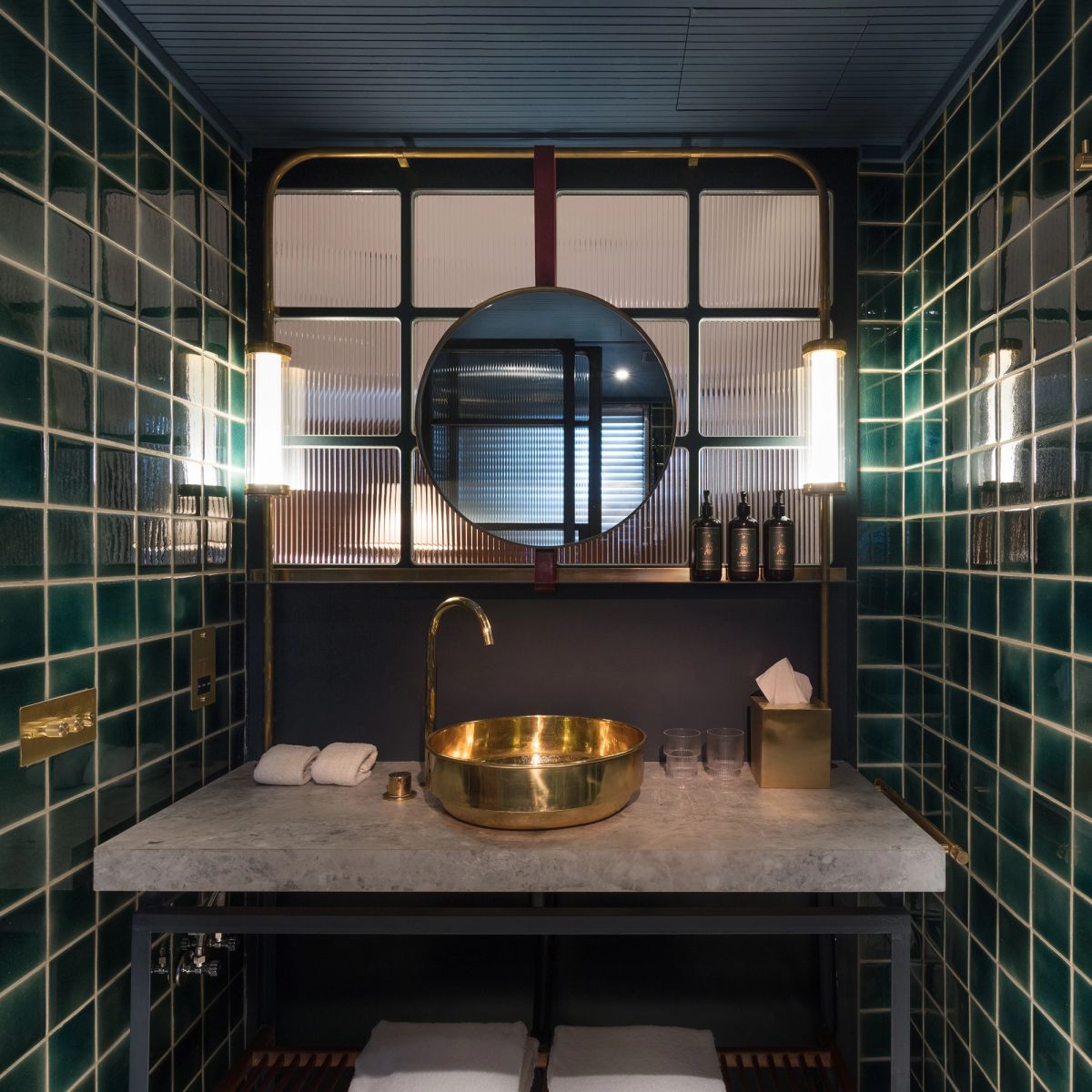A modern bathroom with dark green tiled walls, a concrete vanity, a gold vessel sink, and a round mirror.