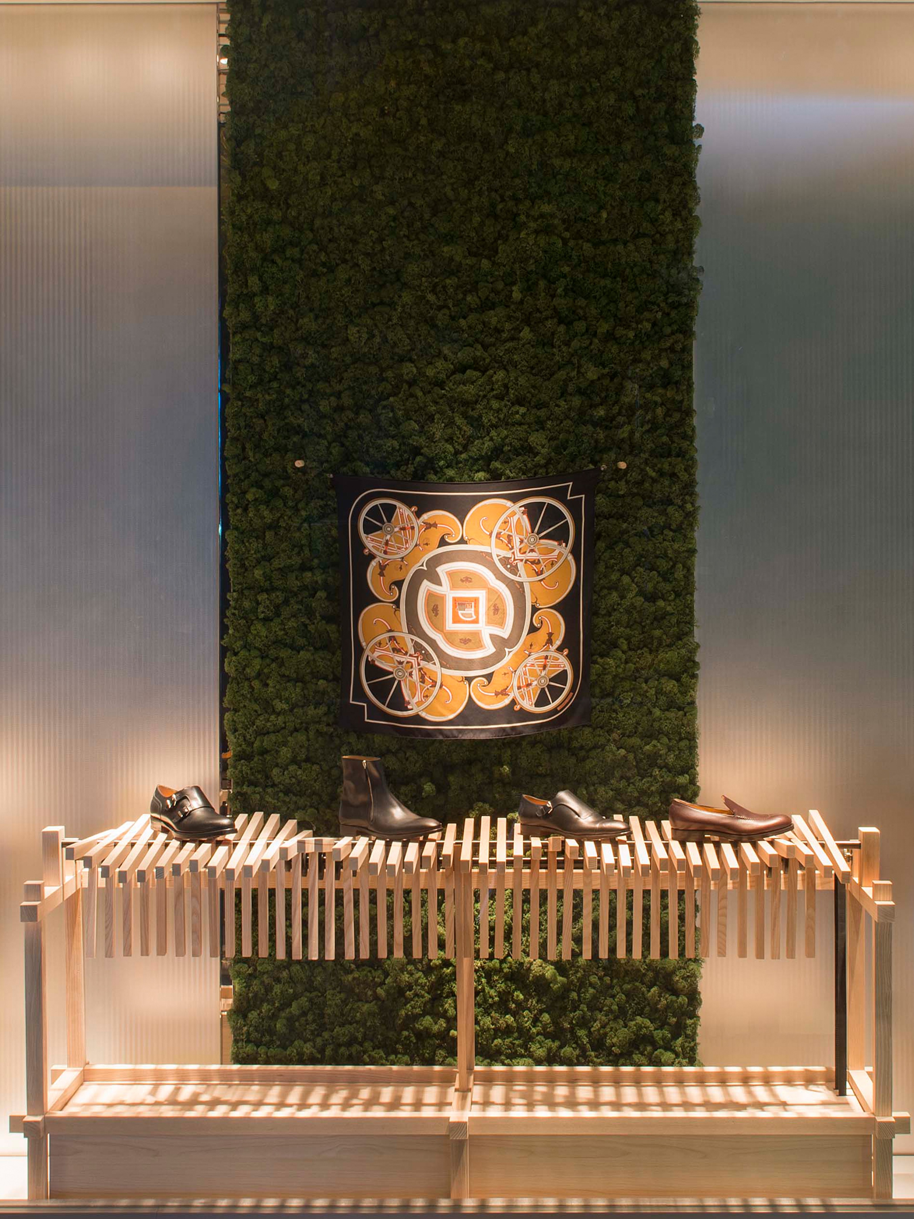 A retail display featuring a green moss wall, a black patterned scarf, and four pairs of men's shoes on a slatted wooden stand.