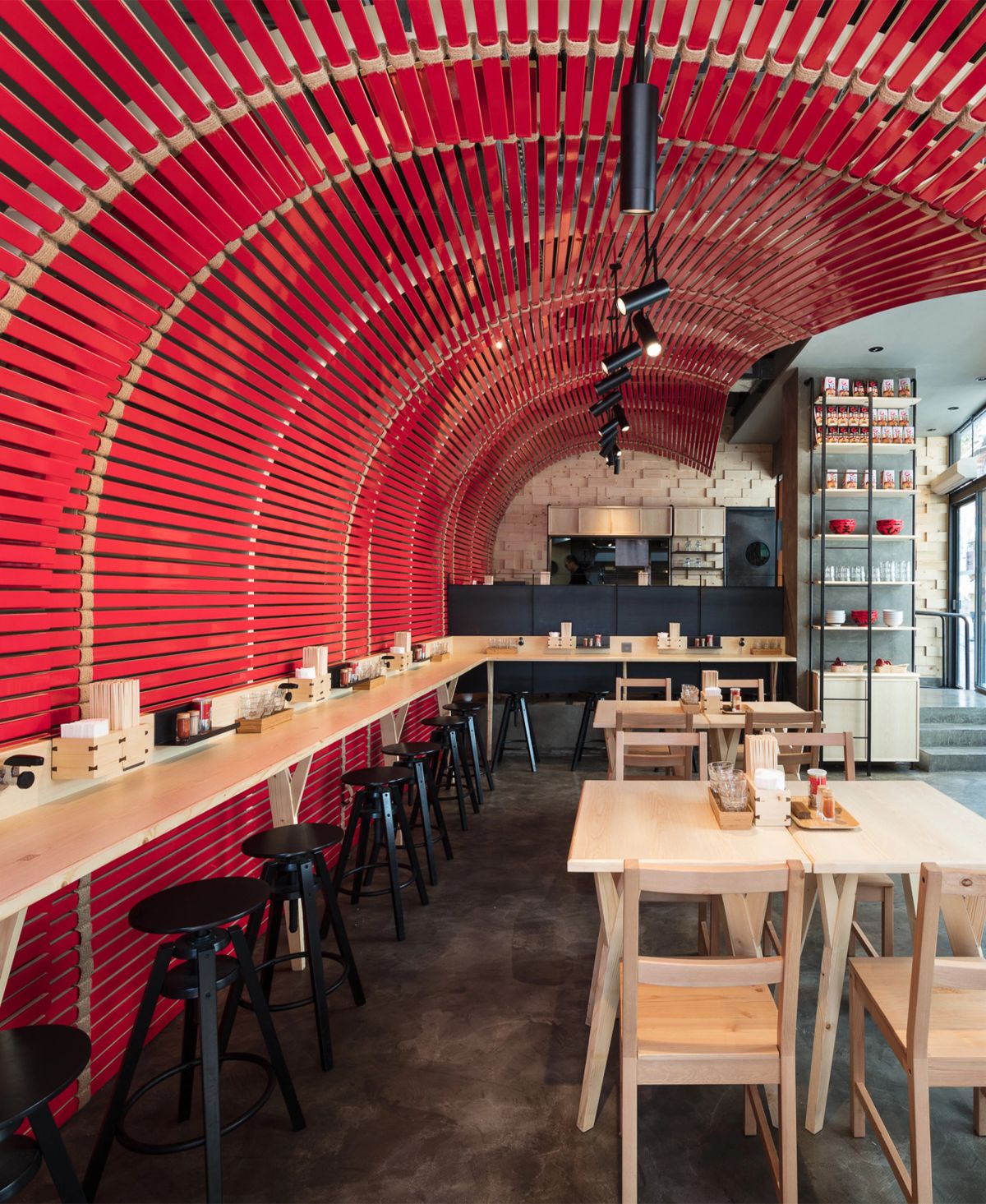 A modern restaurant interior featuring a vibrant red slatted arched ceiling and wall, with a long wooden counter, stools, and light wood tables.