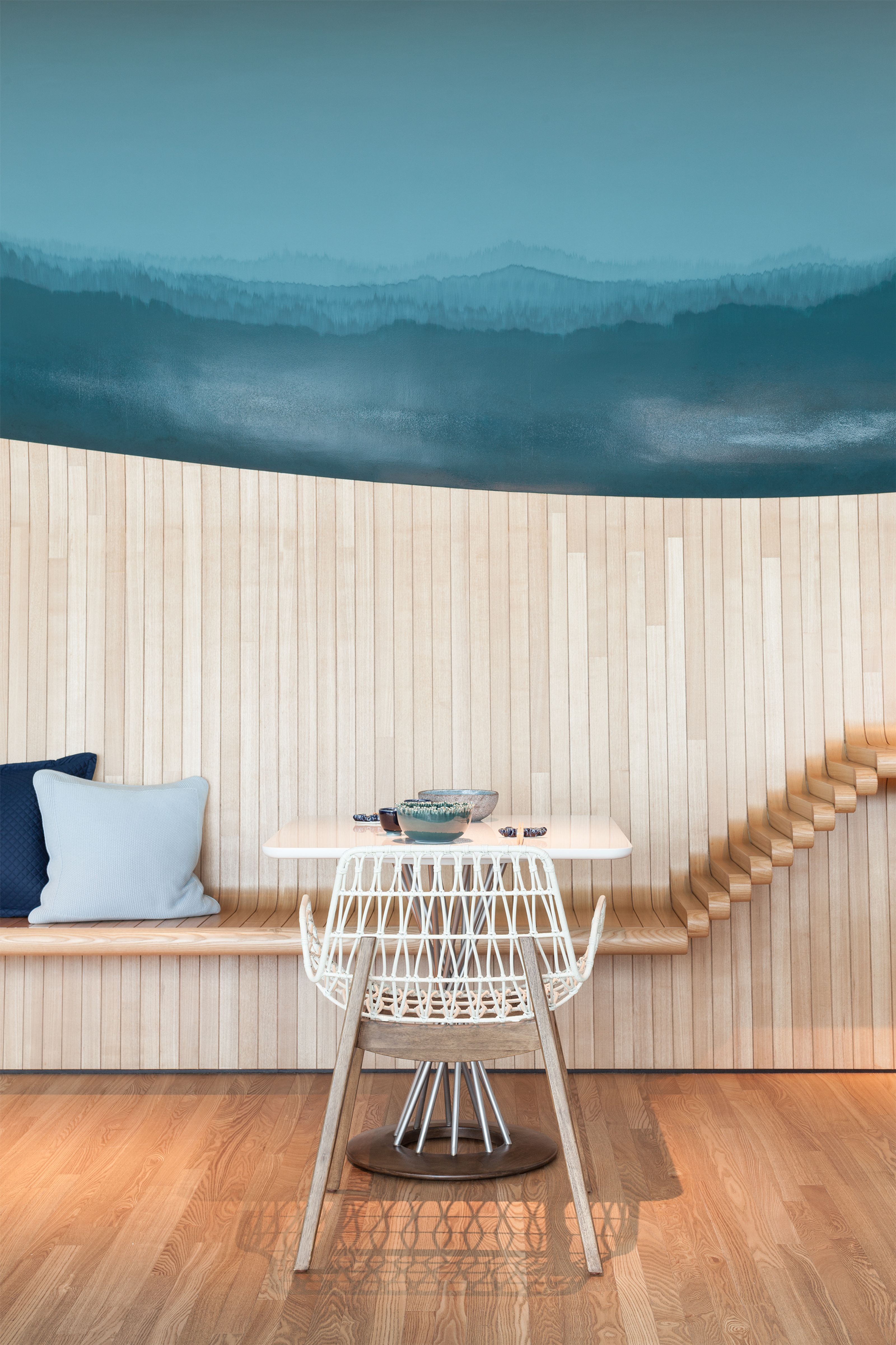 Modern interior with a blue mountain mural above a curved wooden wall and bench, a white table with bowls, a white woven chair, and a sculptural wooden detail.