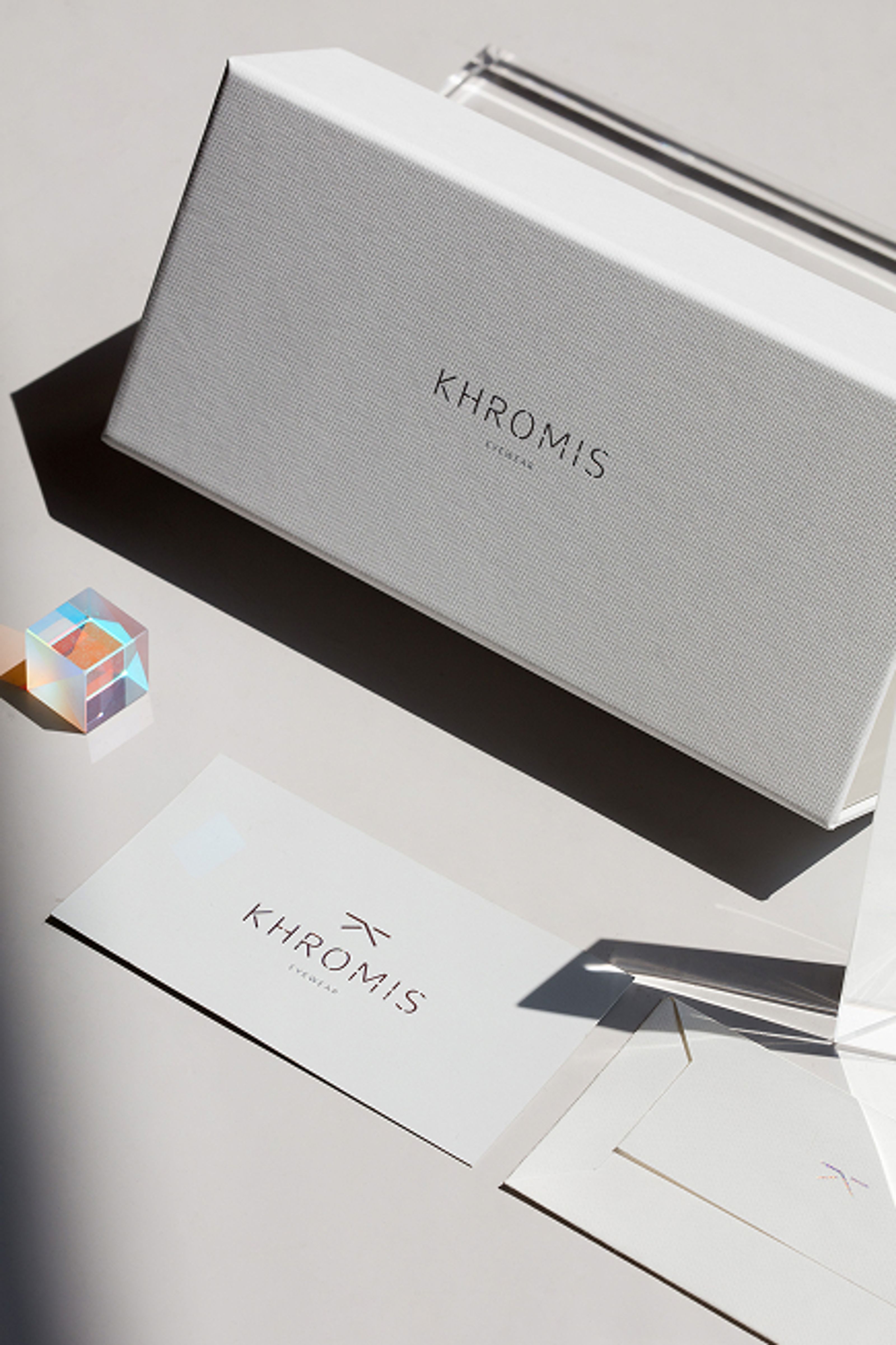A white textured box and a branded card for KHROMIS EYEWEAR, with a small dichroic glass cube, on a light surface with shadows.