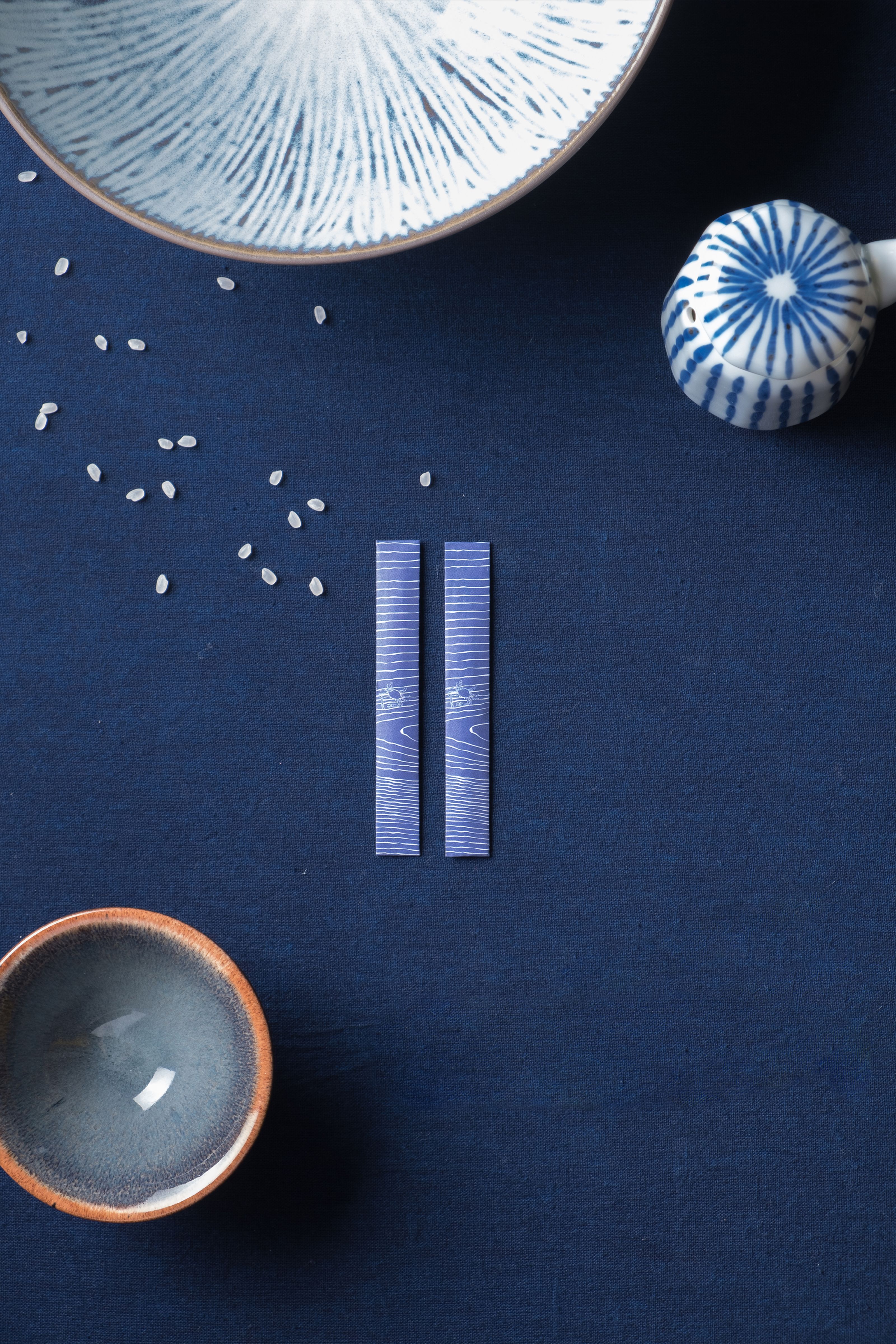 Japanese dining items on a dark blue cloth: blue patterned chopsticks, two ceramic bowls, a small container, and scattered rice.