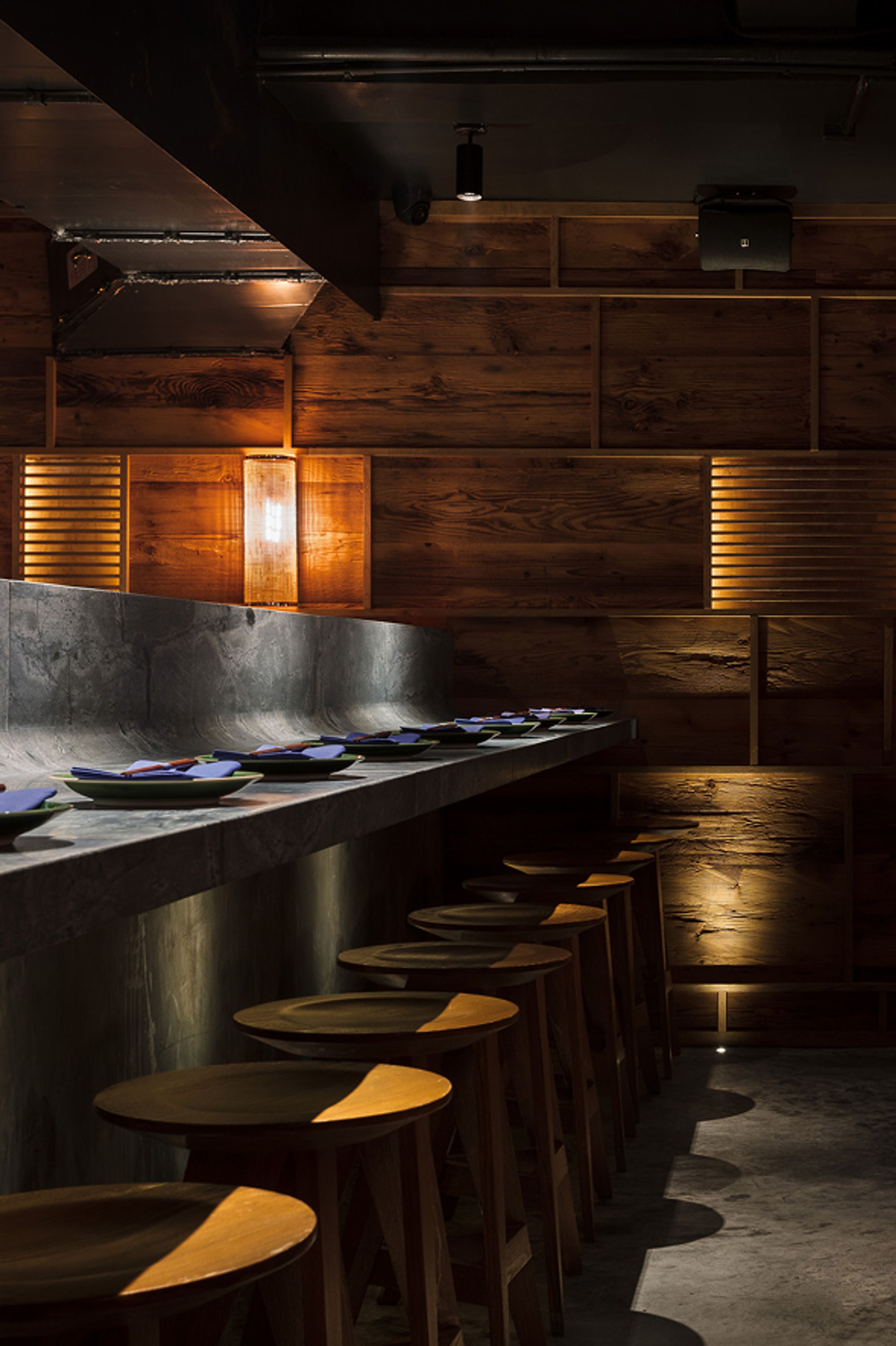 A dimly lit bar features a concrete counter set with plates, a row of wooden stools, and a textured wood-paneled wall.