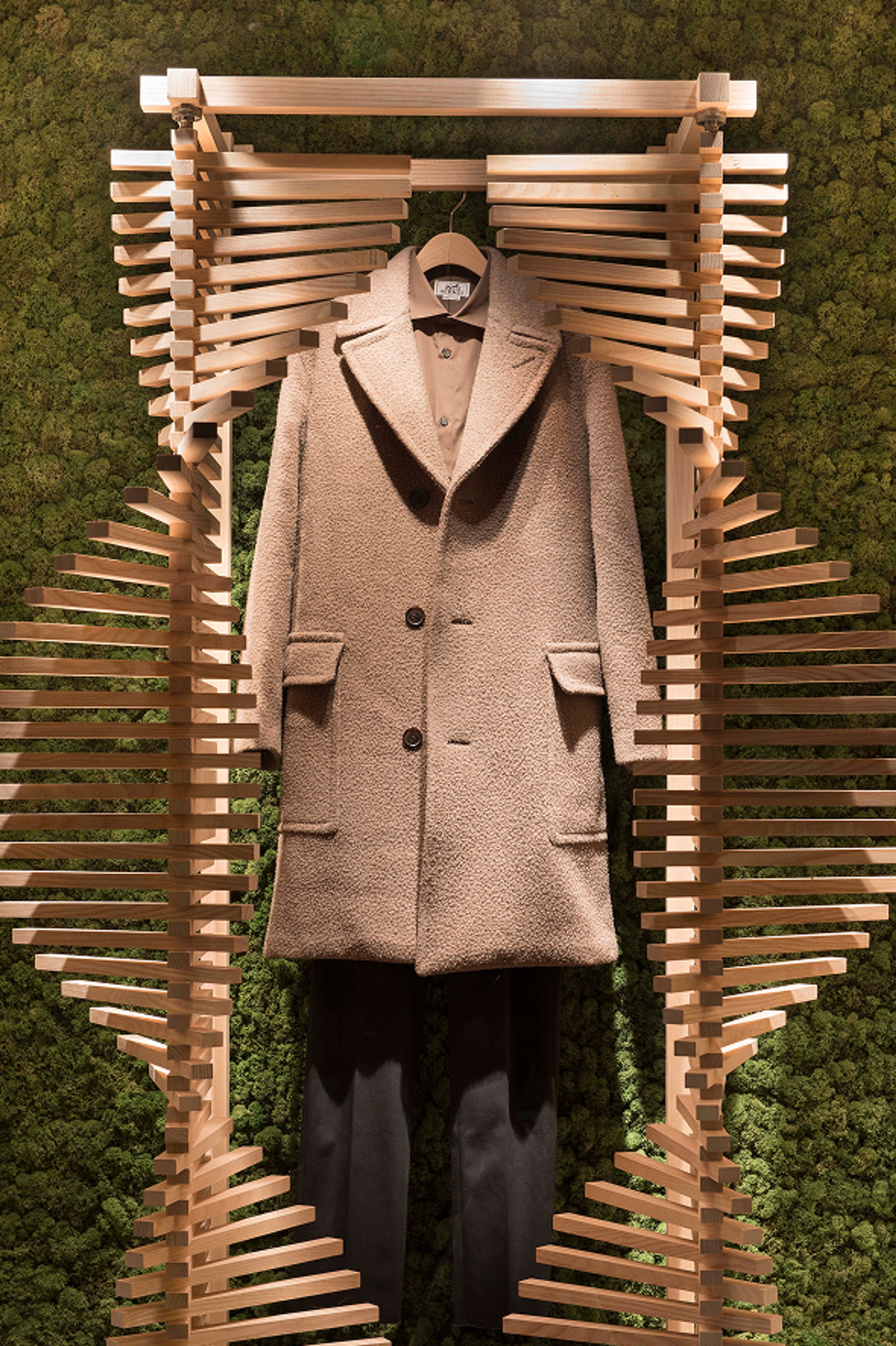A tan textured coat, brown shirt, and dark pants hang from a wooden hanger, framed by decorative wooden slats against a green moss wall.