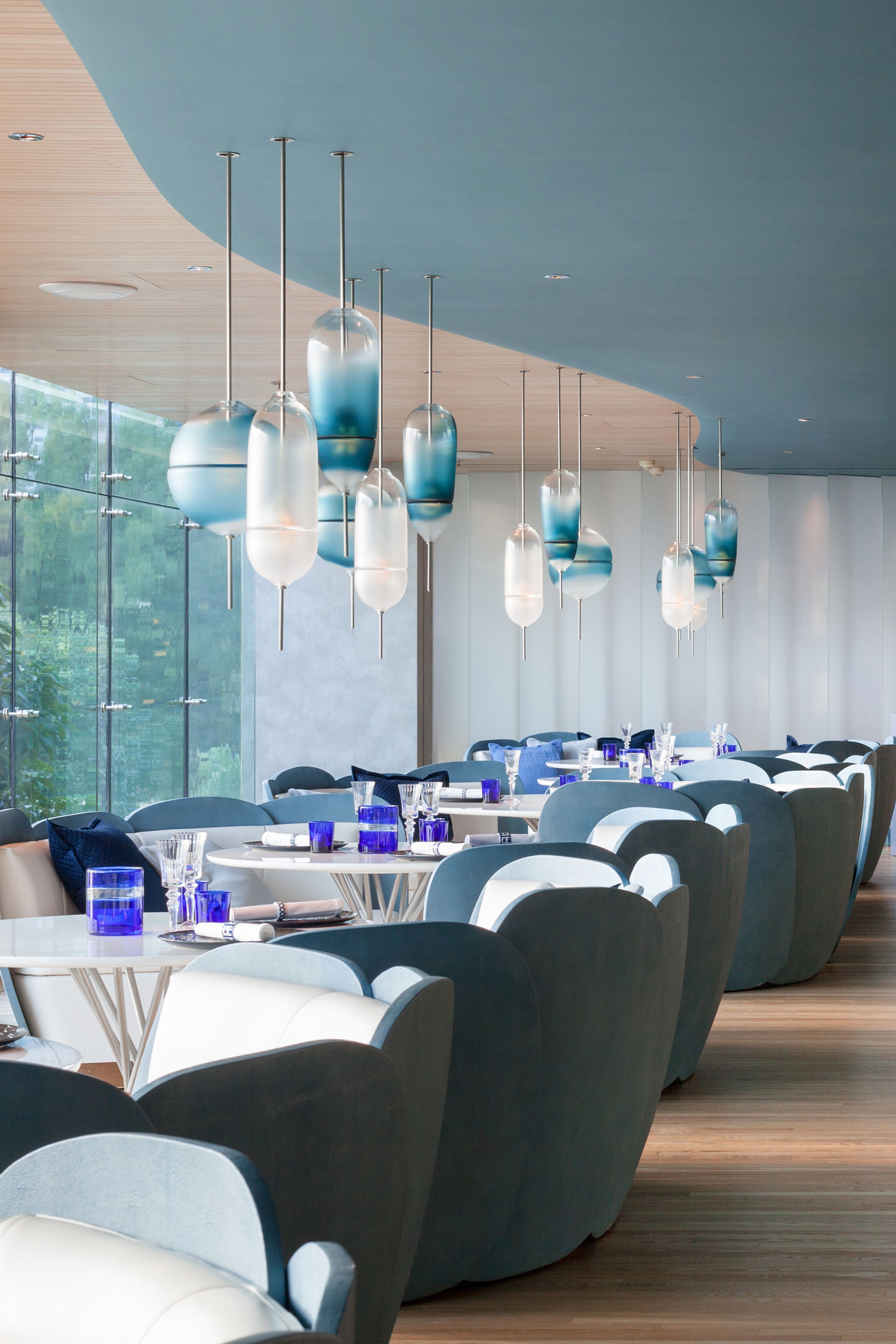 A modern restaurant interior with blue and white decor, featuring ombre glass pendant lights, curved blue chairs, and tables with blue glassware next to a large window.