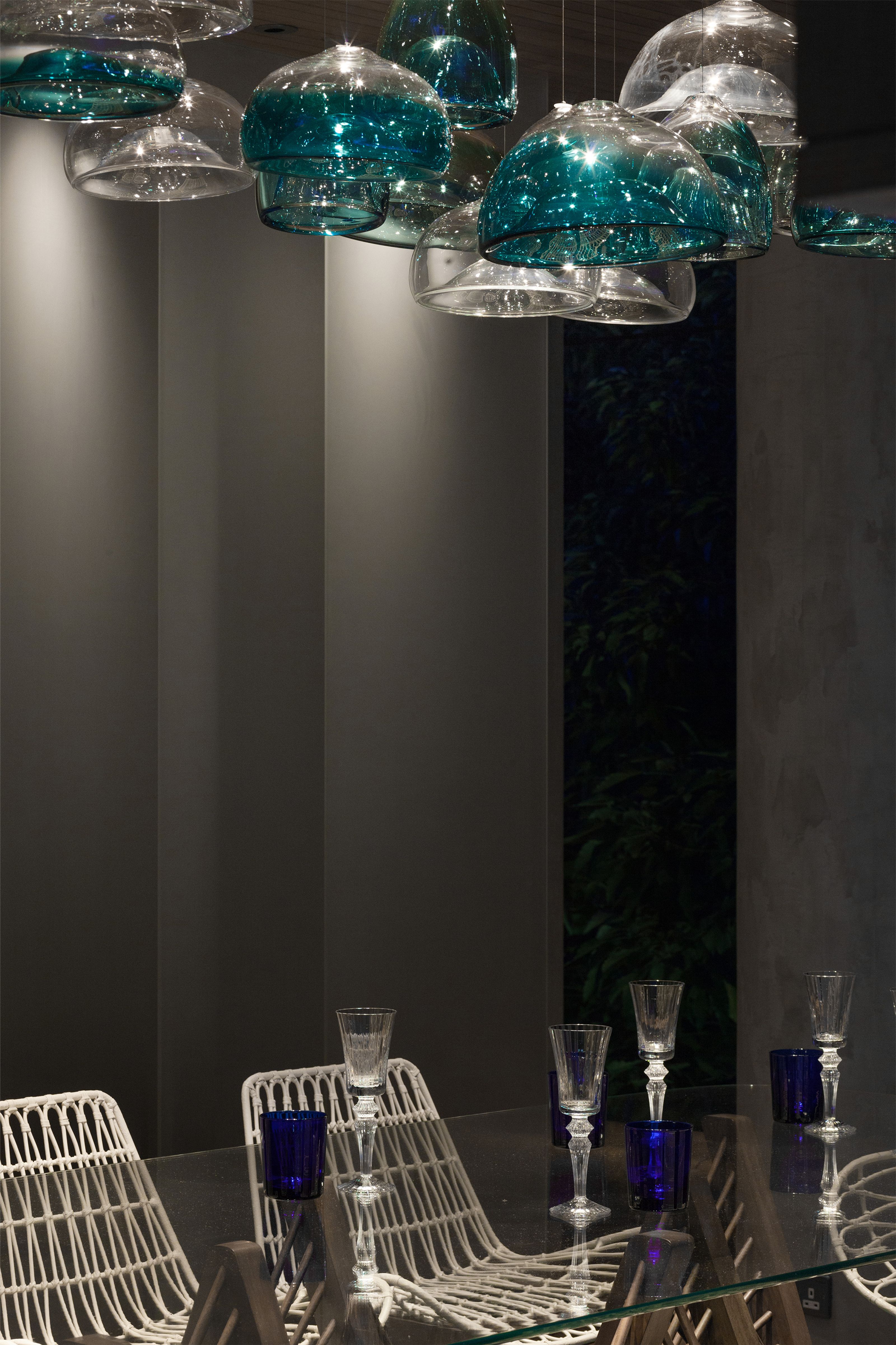 A modern dining room with clear and teal glass pendant lights, a glass table set with white chairs, blue tumblers, and stemmed glasses.