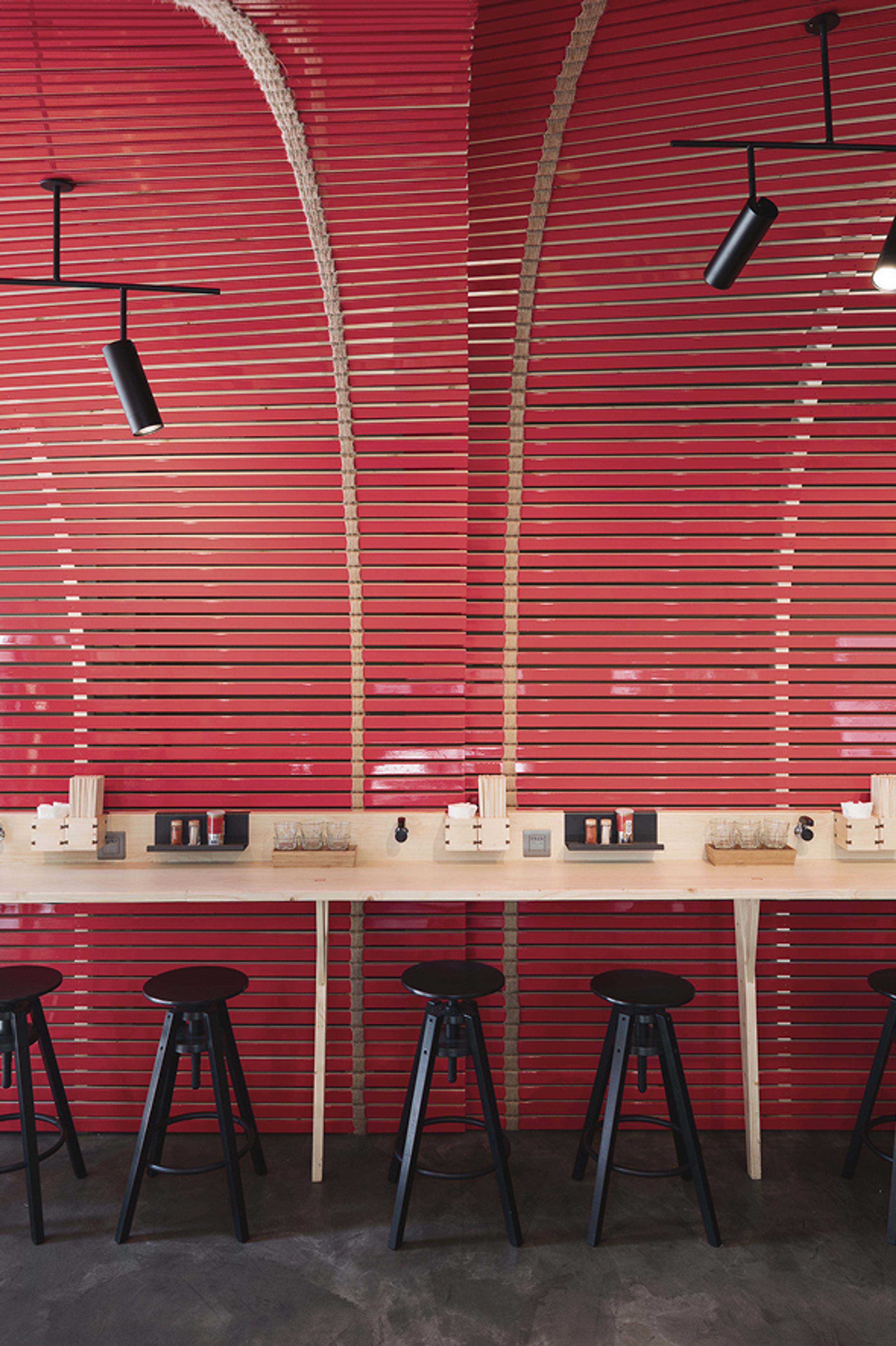 A modern dining space with a red slatted wall curving overhead, a light wooden counter, and black bar stools.