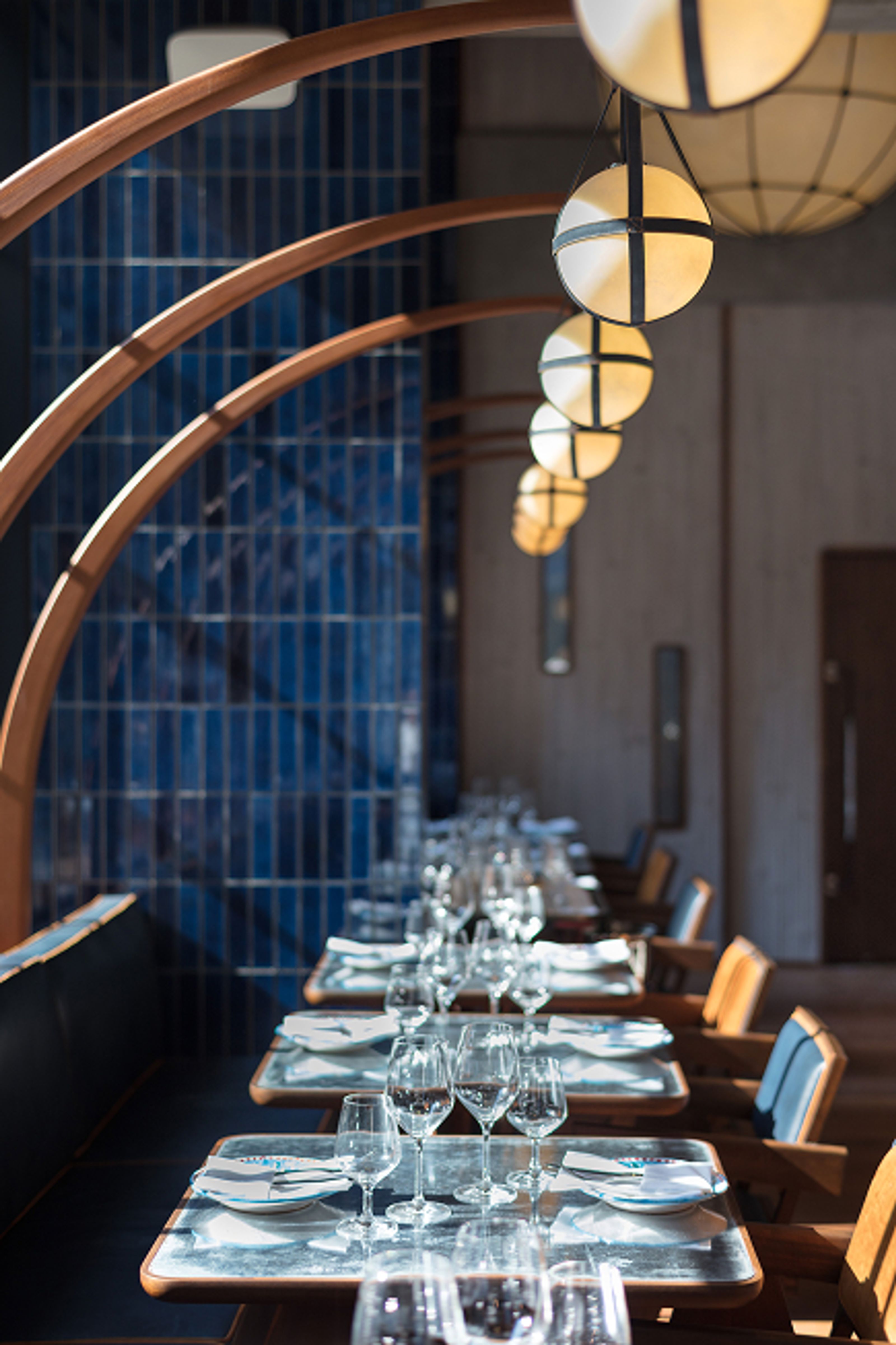 A modern restaurant interior with a row of tables set for dining, blue tiled walls, wooden archways, and hanging spherical pendant lights.