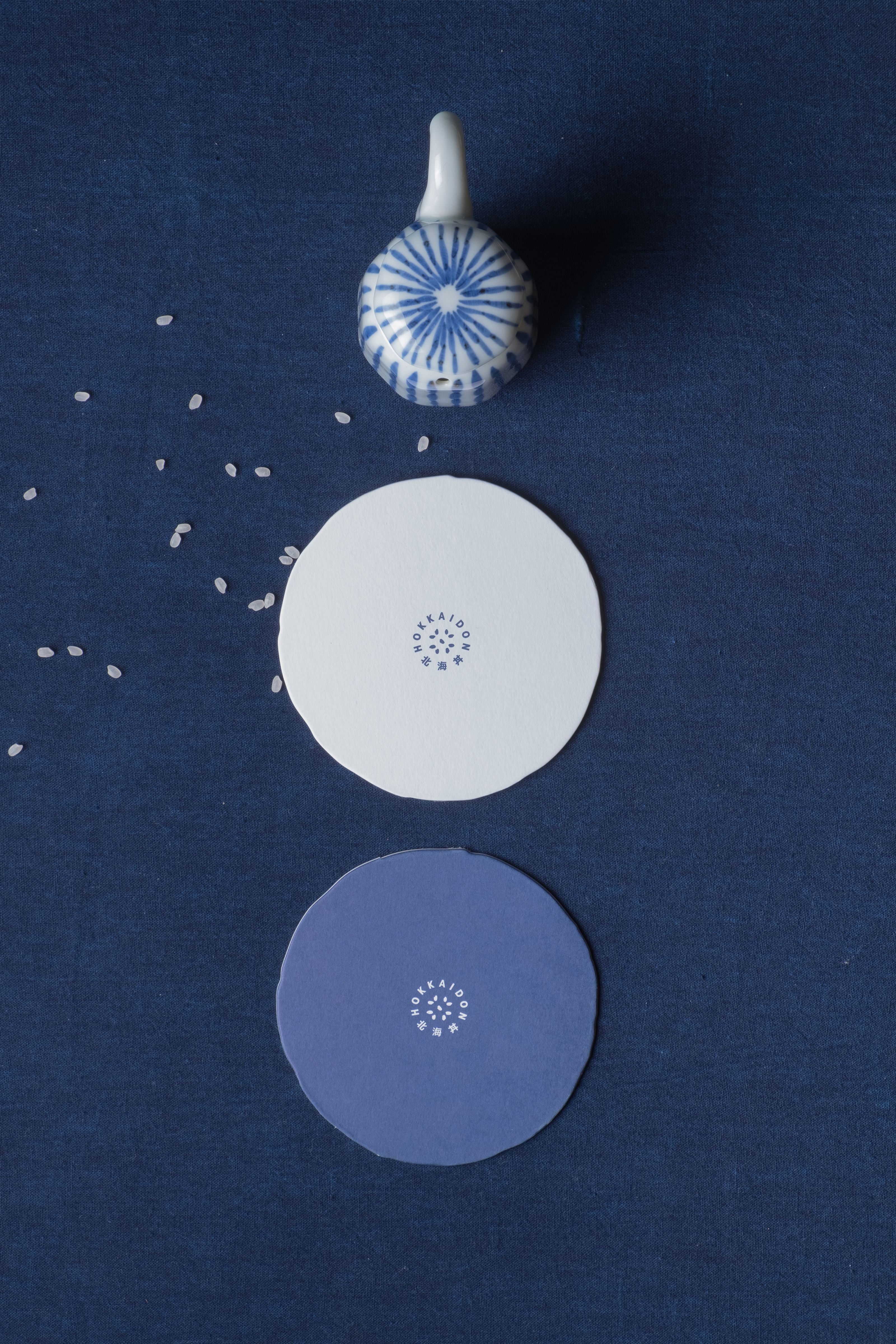 A white and blue ceramic ornament, a white Hokkaido Sake coaster, and a purple Hokkaido Sake coaster are arranged vertically on a dark blue background with scattered rice grains.