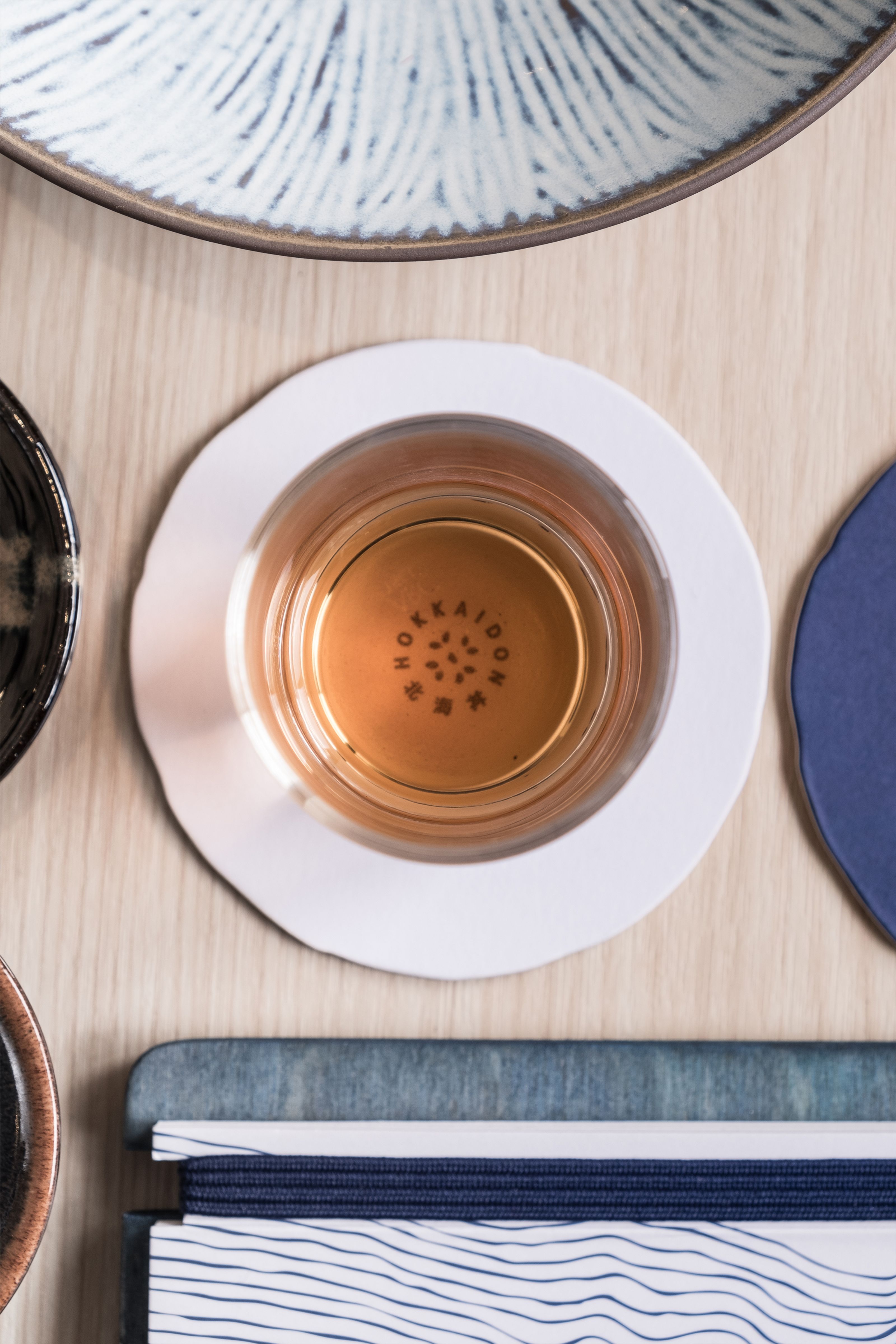 Top-down view of a glass of amber liquid on a white coaster, with "HOKKAIDO" visible at the bottom, surrounded by patterned dishes and notebooks on a light wooden table.