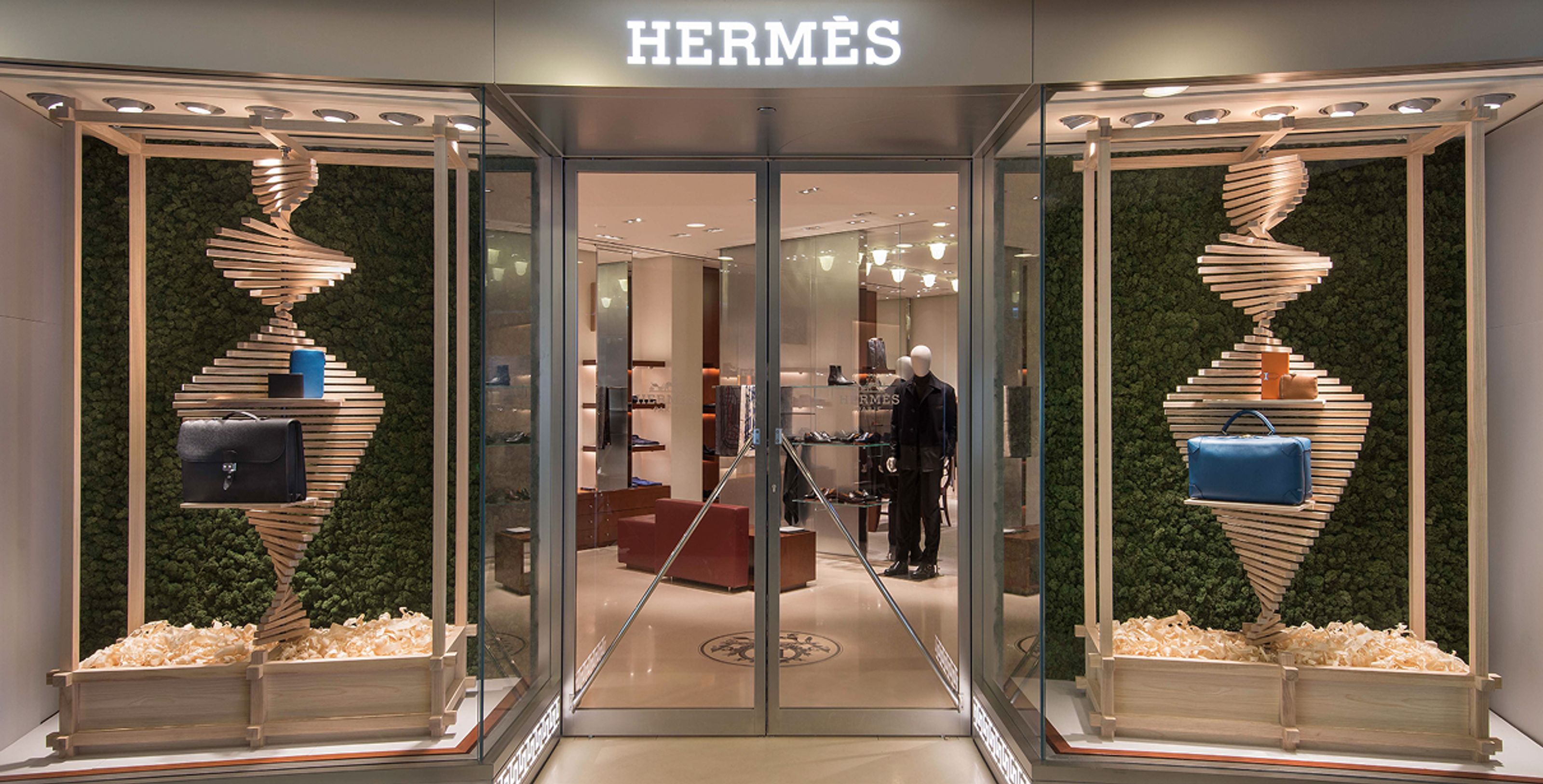 Hermès store entrance flanked by display windows featuring spiral wooden shelves against a moss wall, showcasing handbags.