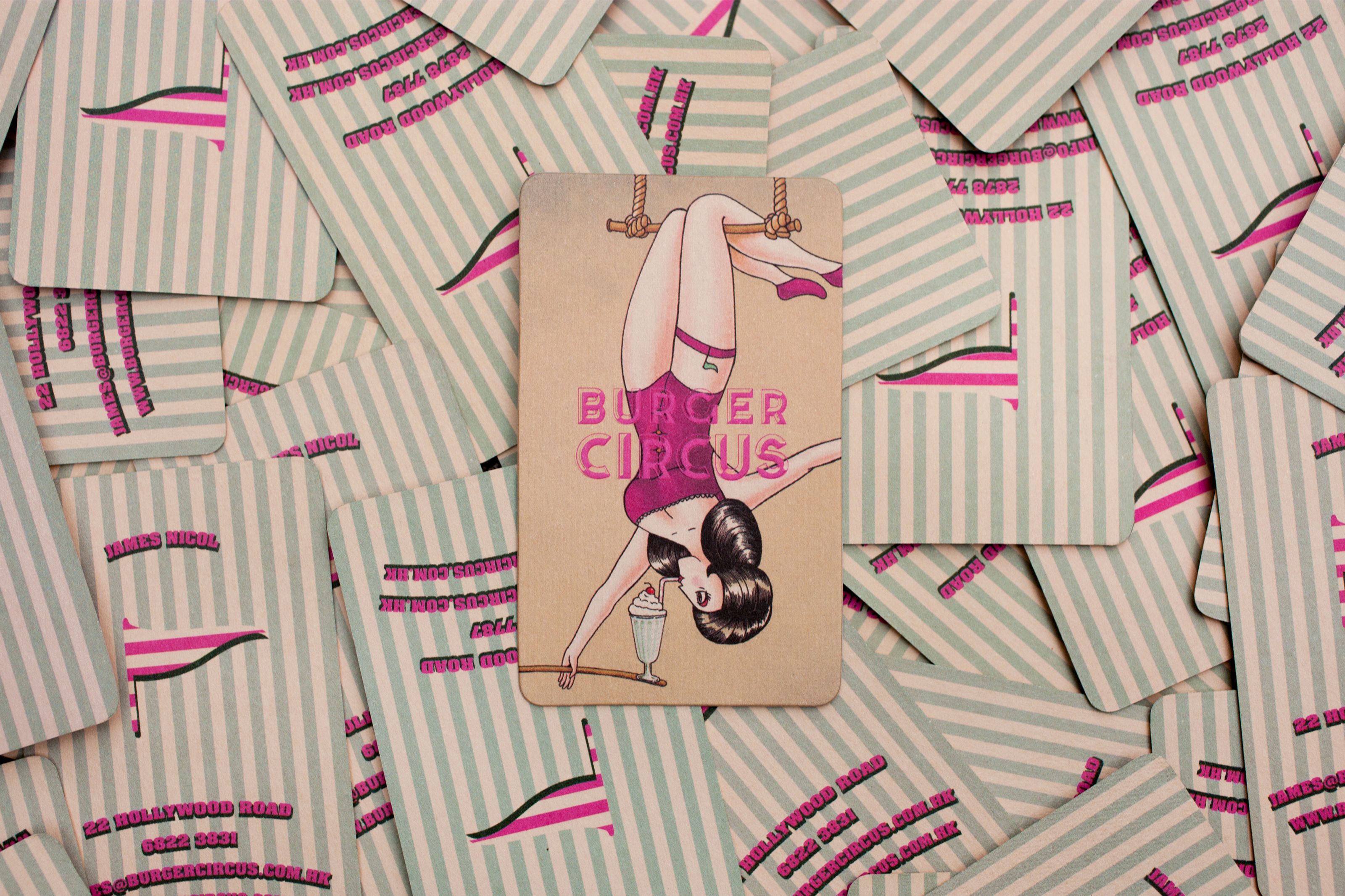 A Burger Circus business card shows a pin-up woman hanging upside down from a trapeze, holding a milkshake, on a pile of striped cards.