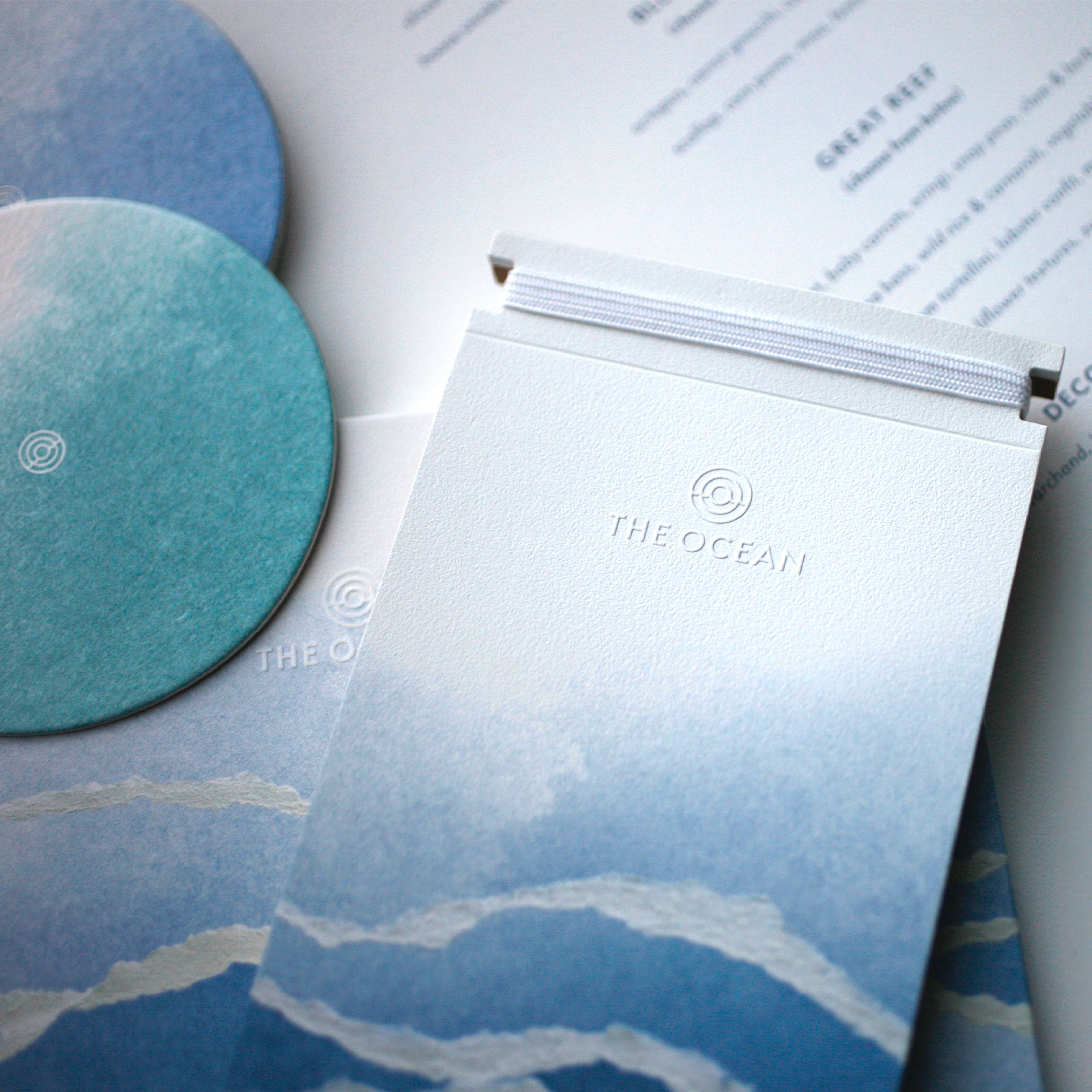 "The Ocean" branded stationery with blue and teal watercolor designs, including a notepad and coasters.