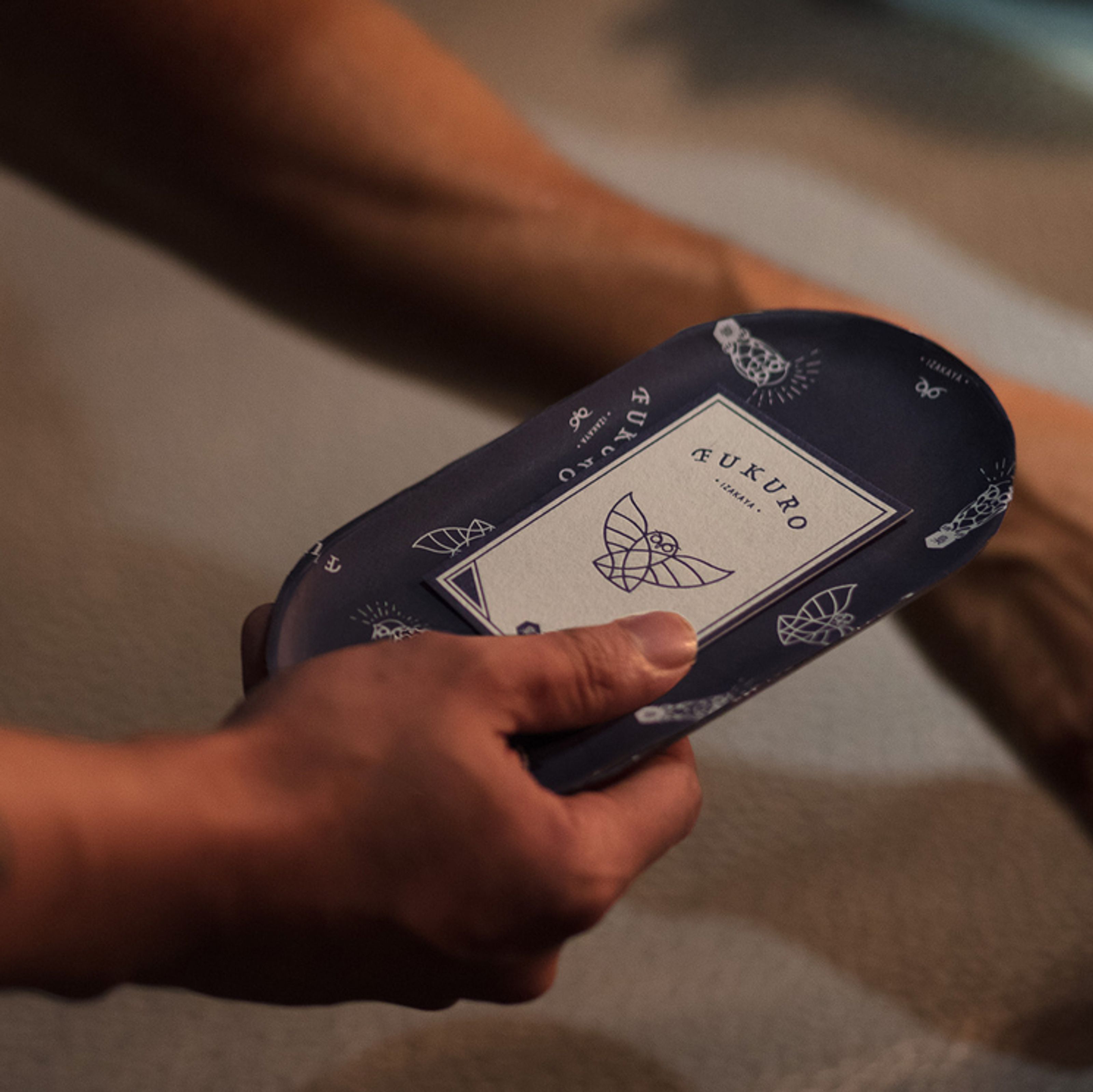 A hand holds a dark blue, oval tray with an owl logo and "FUKURO IZAKAYA" text on a white label.