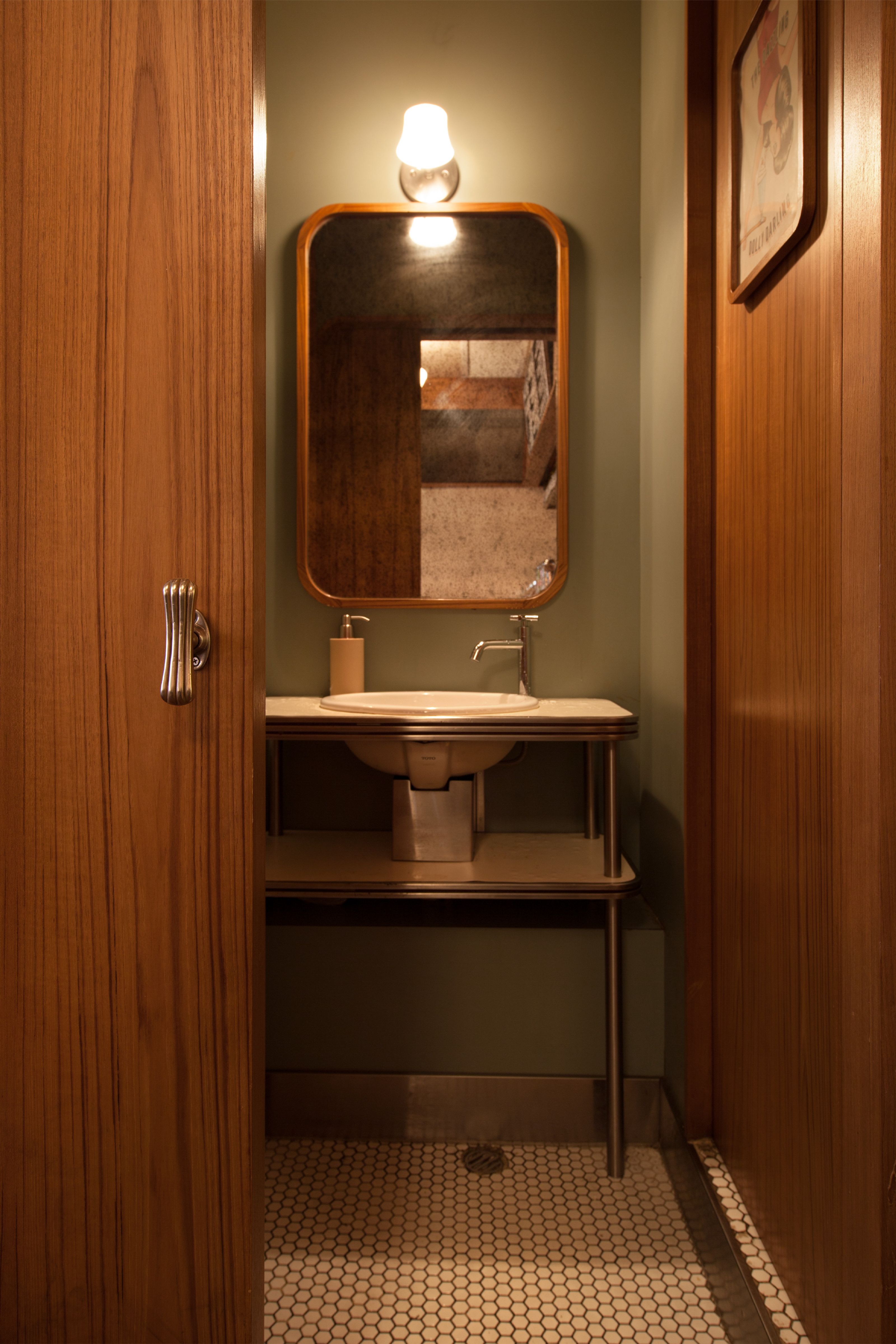 A stylish powder room with olive green walls, a wooden mirror above a sink on a metal stand, and white hexagonal tile flooring.