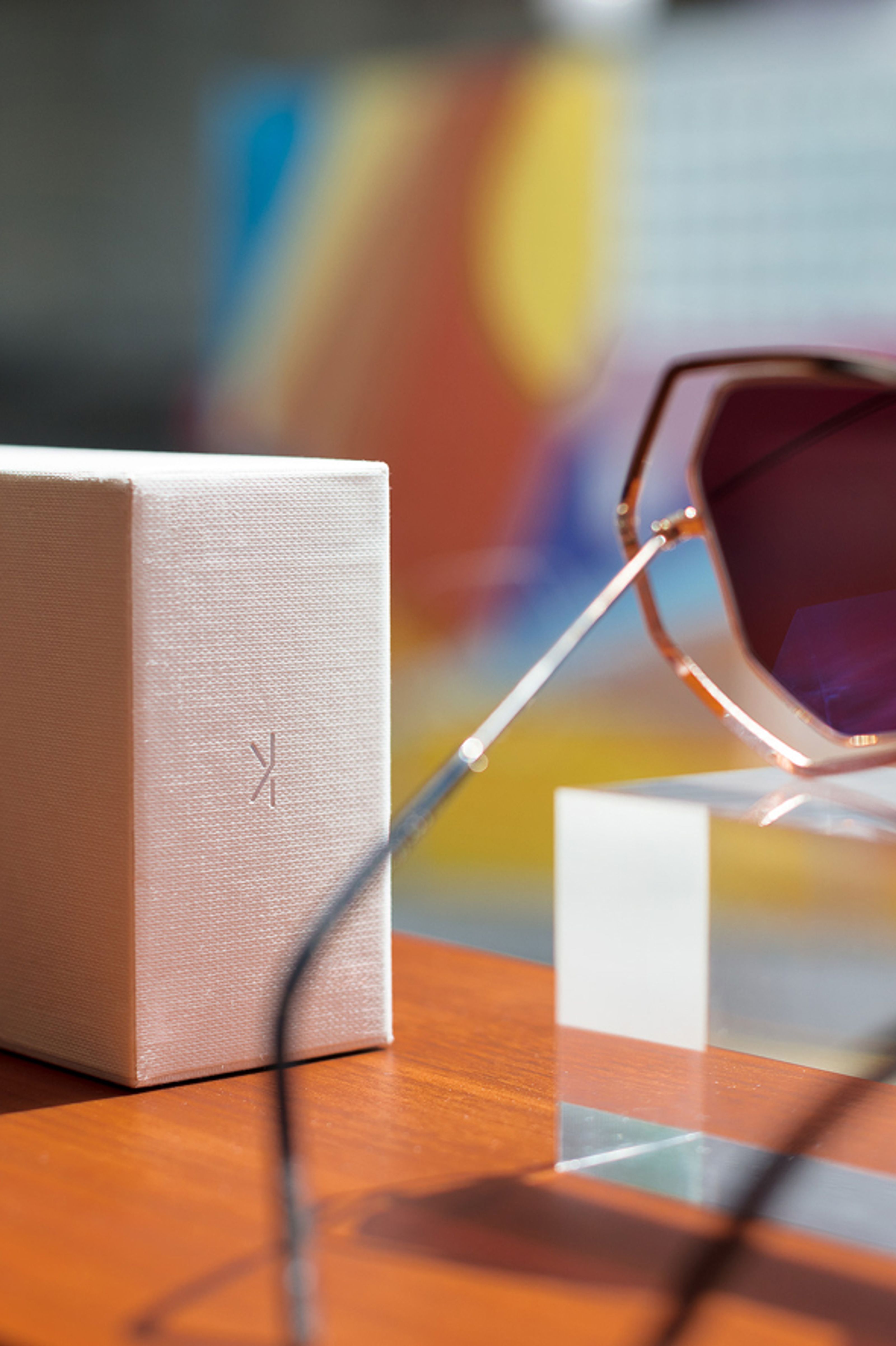 A white textured box with a stylized K logo stands beside rose gold sunglasses on a clear block, all on a wooden surface with a colorful blurred background.