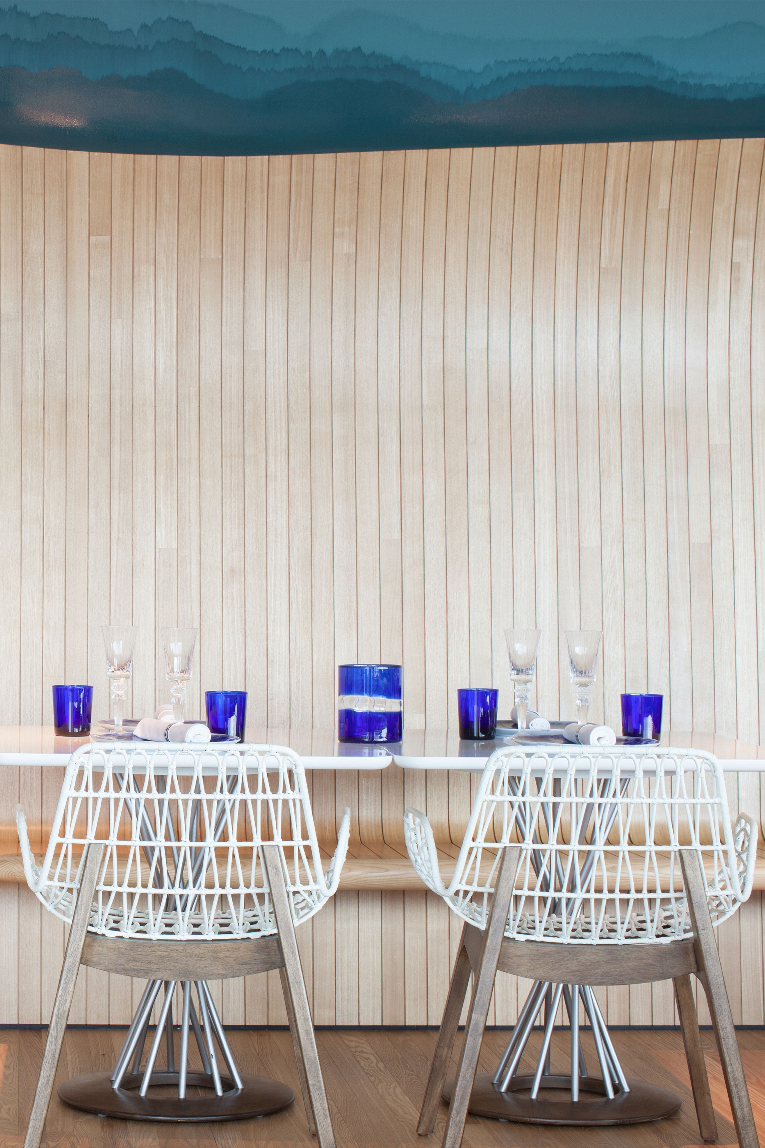 A modern dining room features a blue mountain mural above a light wood slatted wall, a white table set with blue glasses, and white woven chairs.