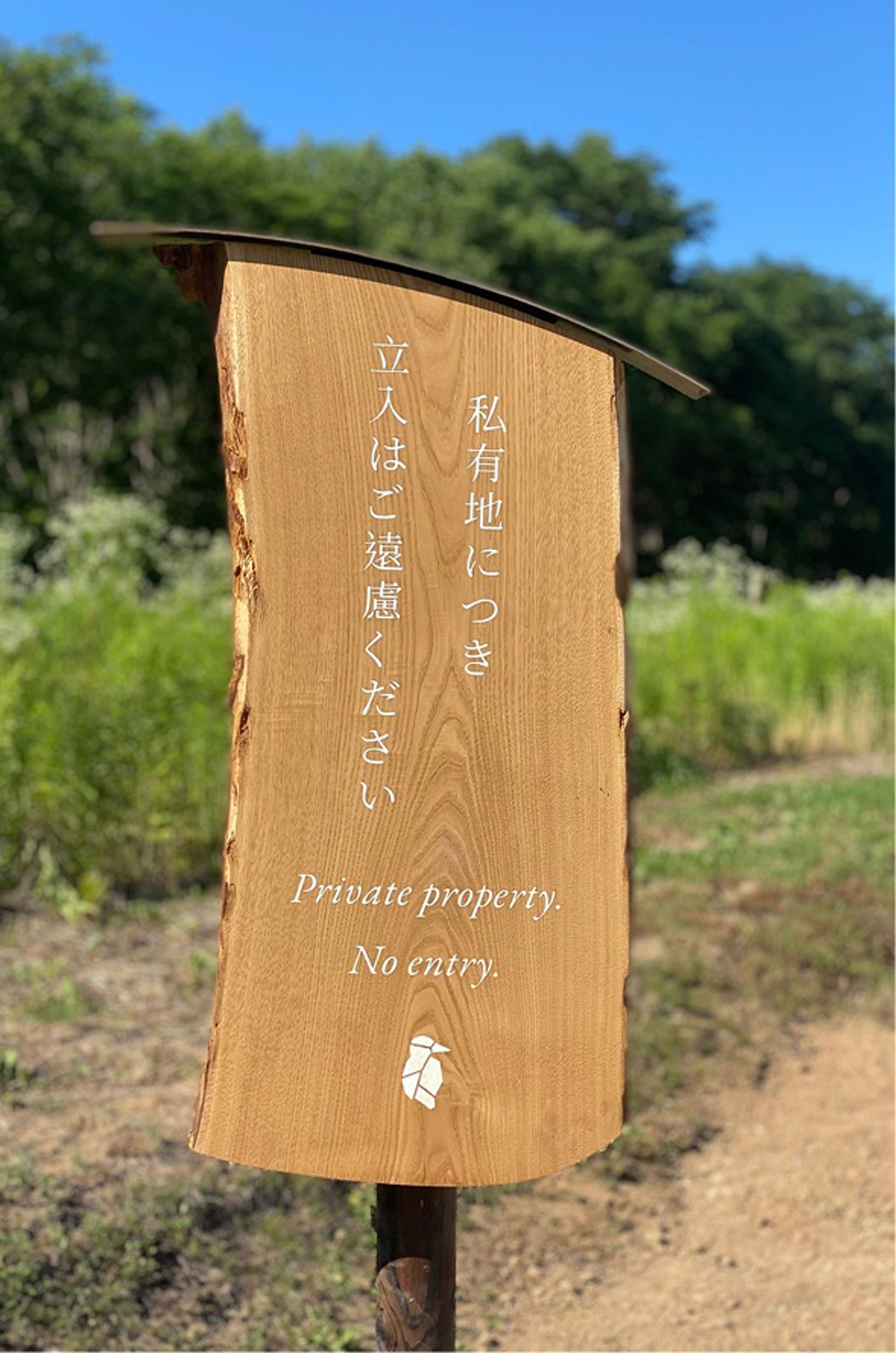 A live-edge wooden sign reads "Private property. No entry." in English and Japanese, with a small bird logo, in a sunny outdoor setting.