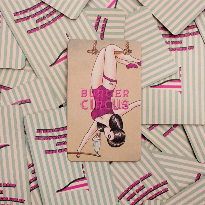 A "Burger Circus" business card featuring a pin-up style woman on a trapeze drinking a milkshake, resting on a pile of striped cards.