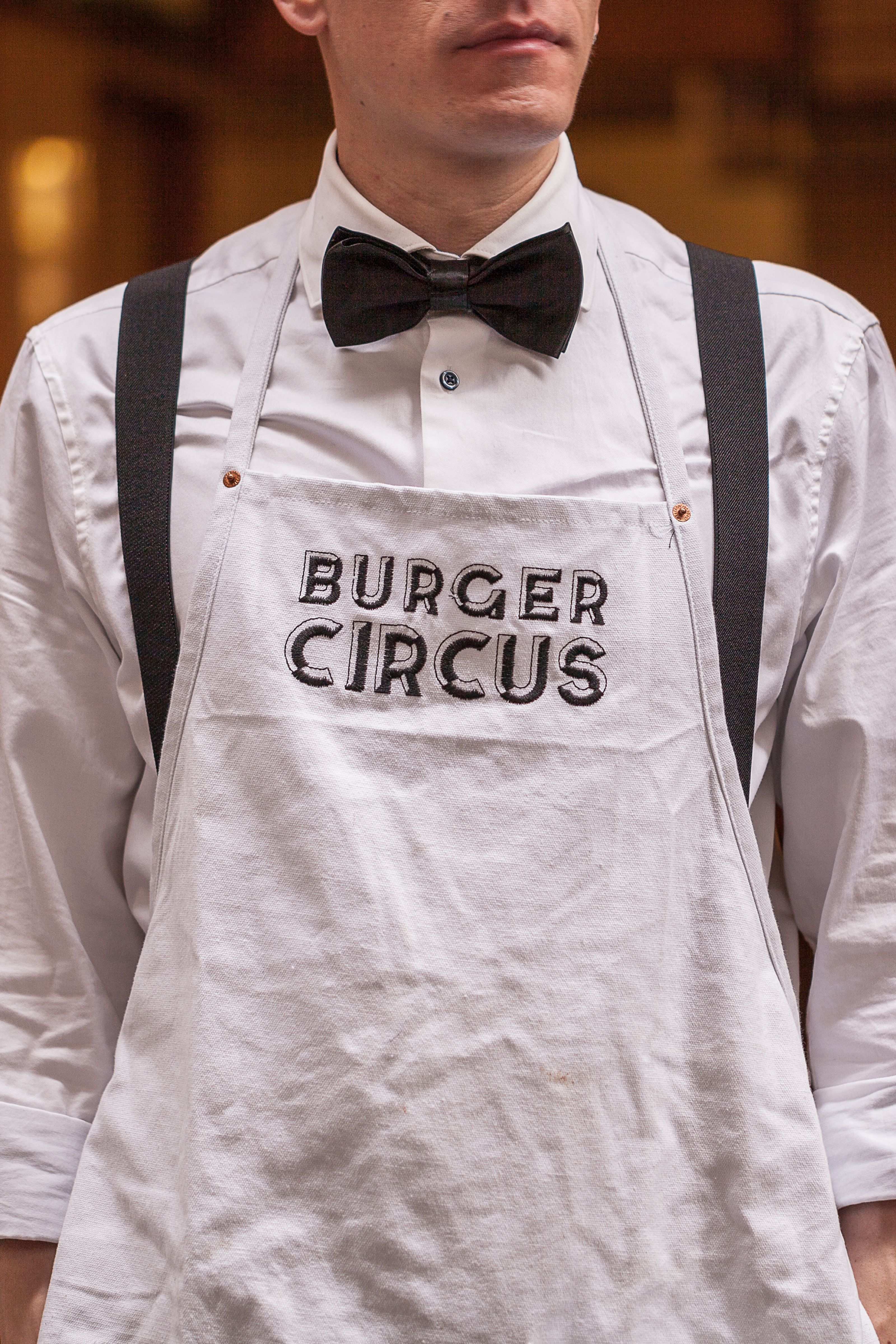 Person wearing a white shirt, black bow tie, suspenders, and a white apron embroidered with "BURGER CIRCUS."