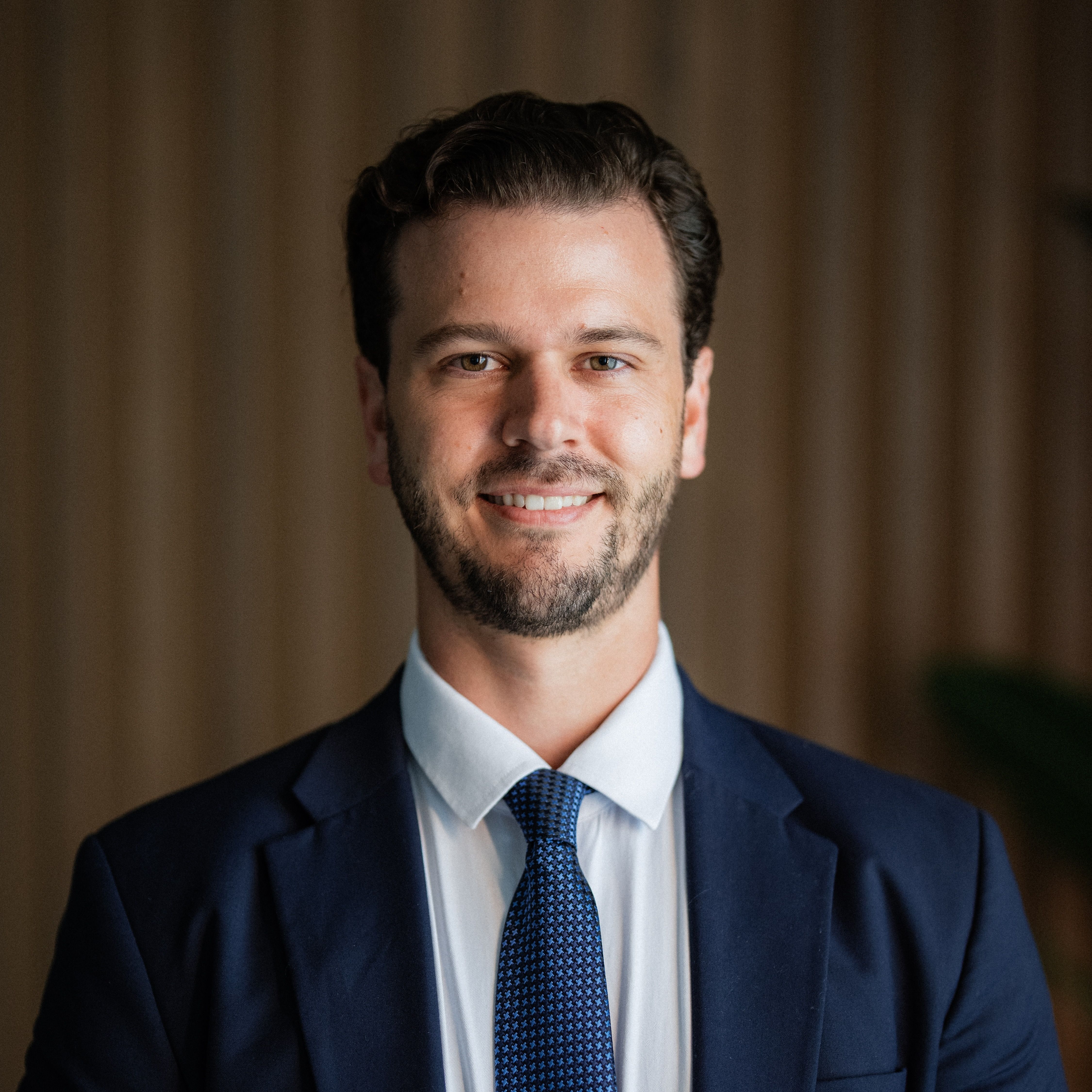 Smiling man with short brown hair and a beard, dressed in a navy blue suit, white shirt, and blue tie.