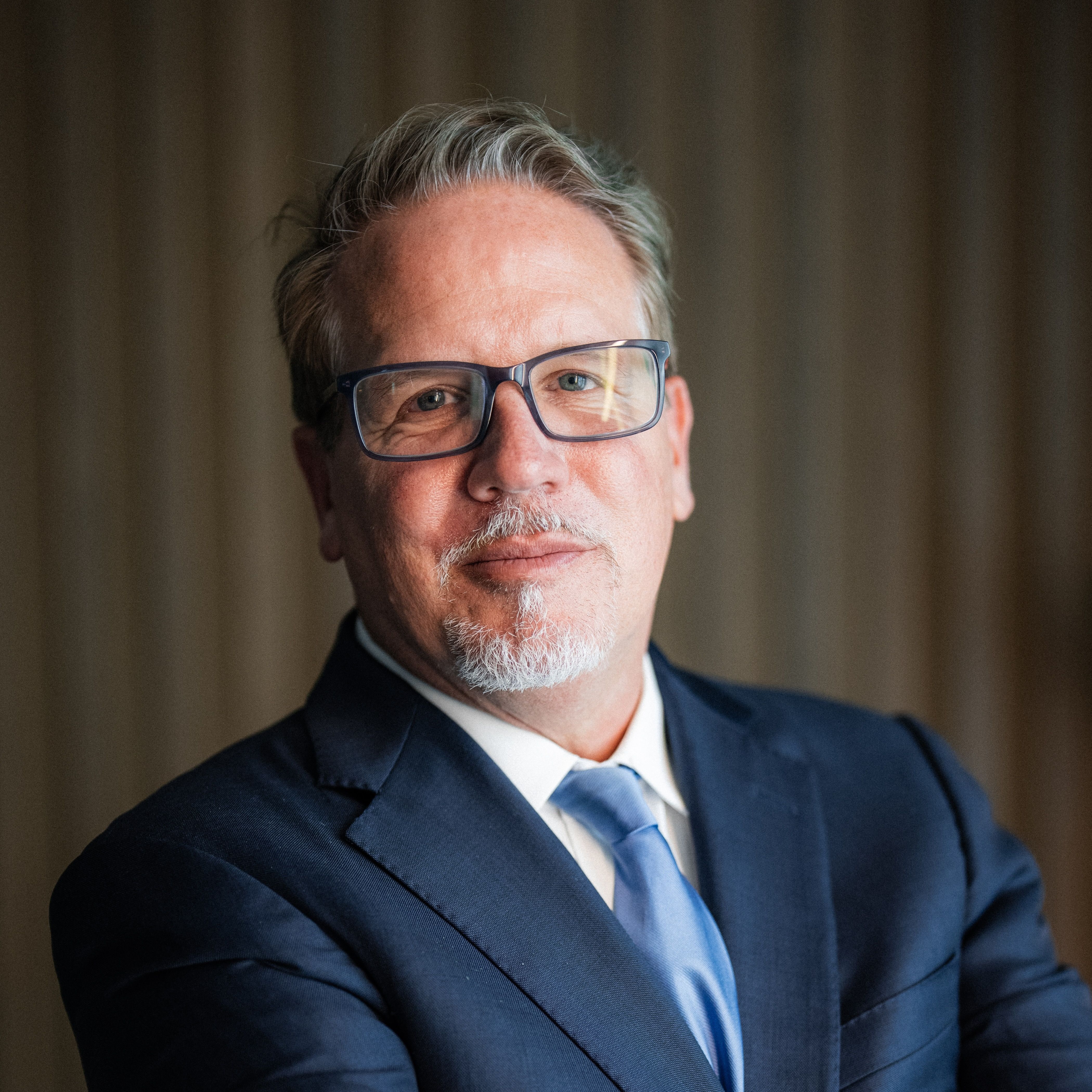 Middle-aged man with glasses and a gray goatee in a navy suit and light blue tie.