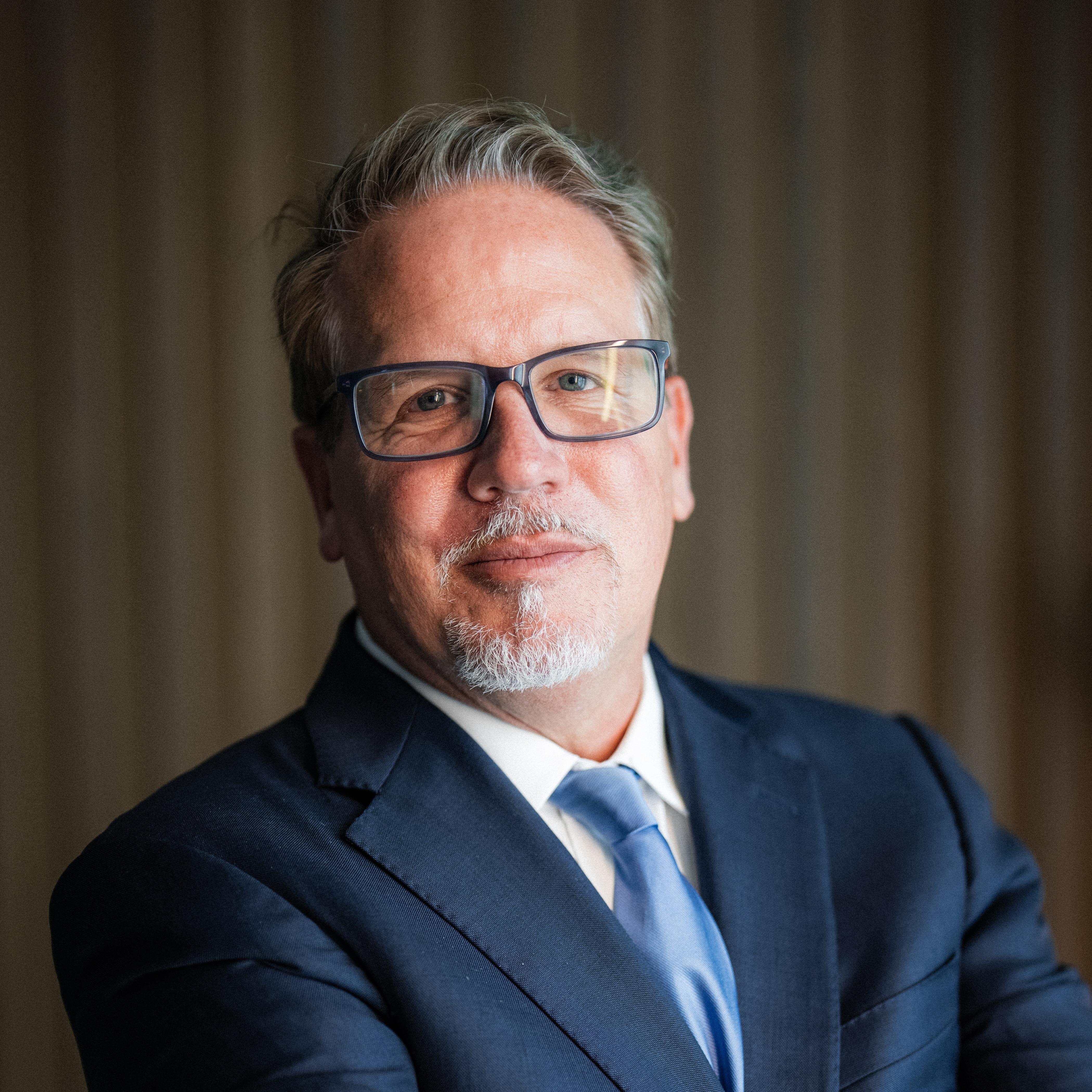 Middle-aged man with glasses and a gray goatee in a navy suit and light blue tie.