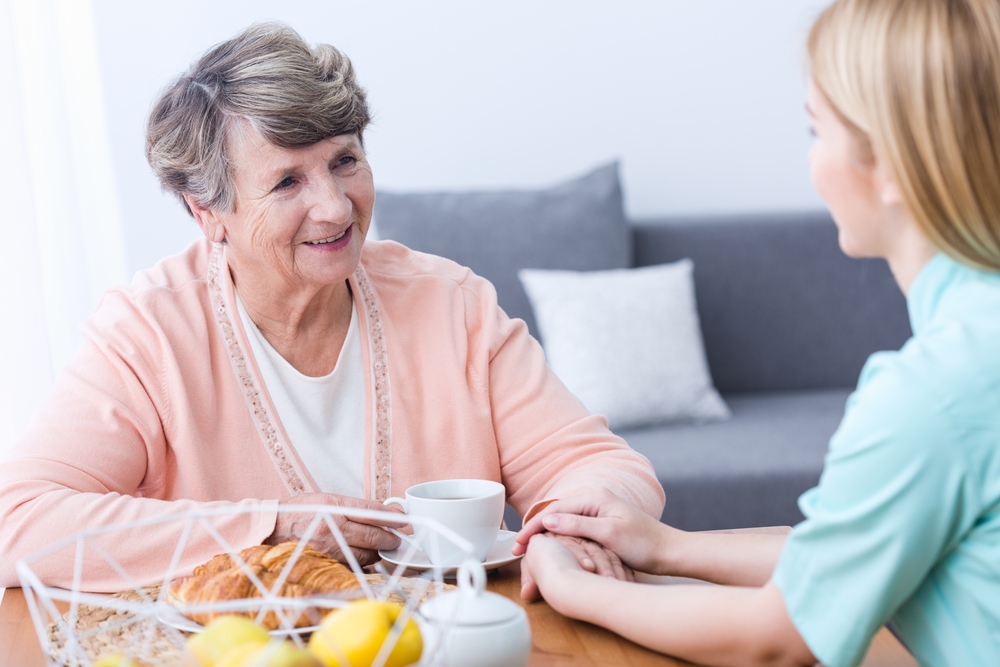 Image A smiling senior woman in a nursing home holding a cup of tea while a caregiver gently holds her hand during a New Year’s visit.
