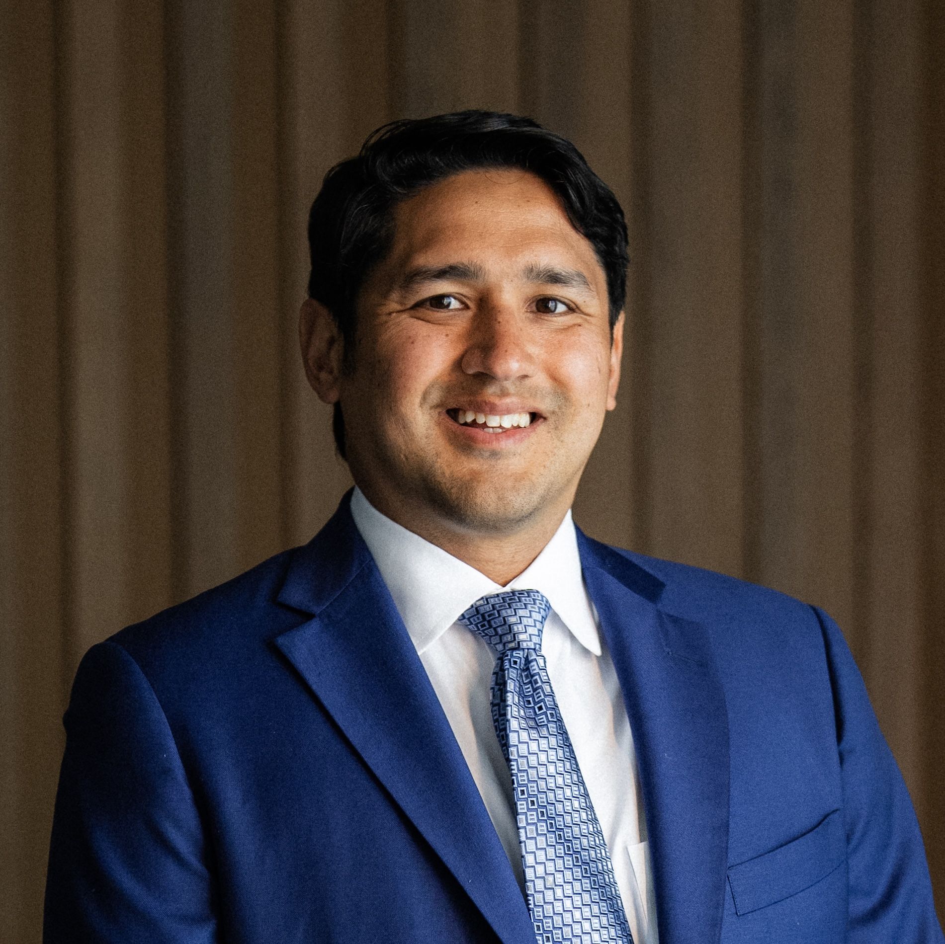 Smiling man in a royal blue suit jacket, white shirt, and patterned blue tie against a neutral background.