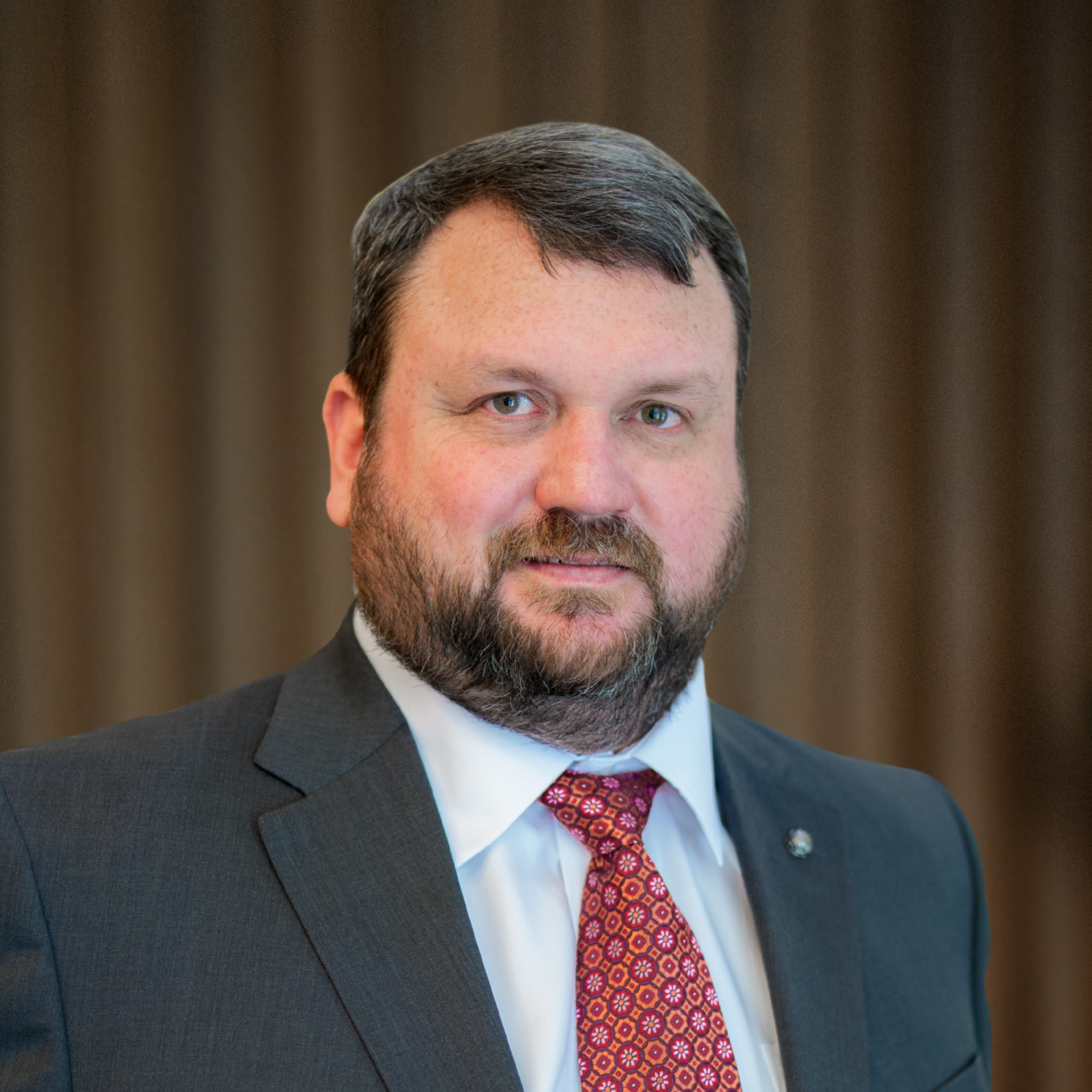 Bearded man in a dark gray suit, white dress shirt, and red patterned necktie smiling against a brown backdrop.