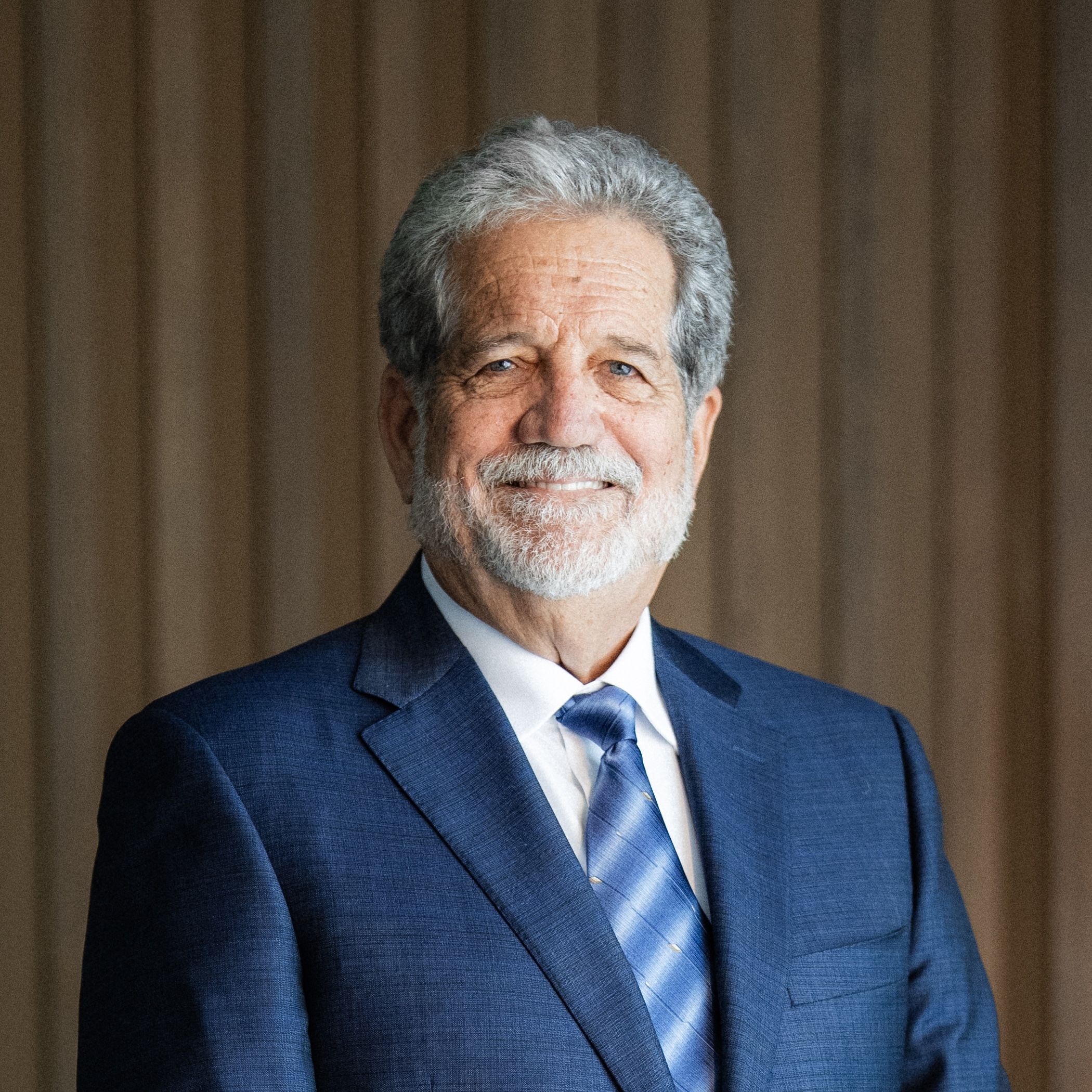 Smiling older man with gray hair and beard wearing a navy blue suit and patterned blue tie against wood paneling.