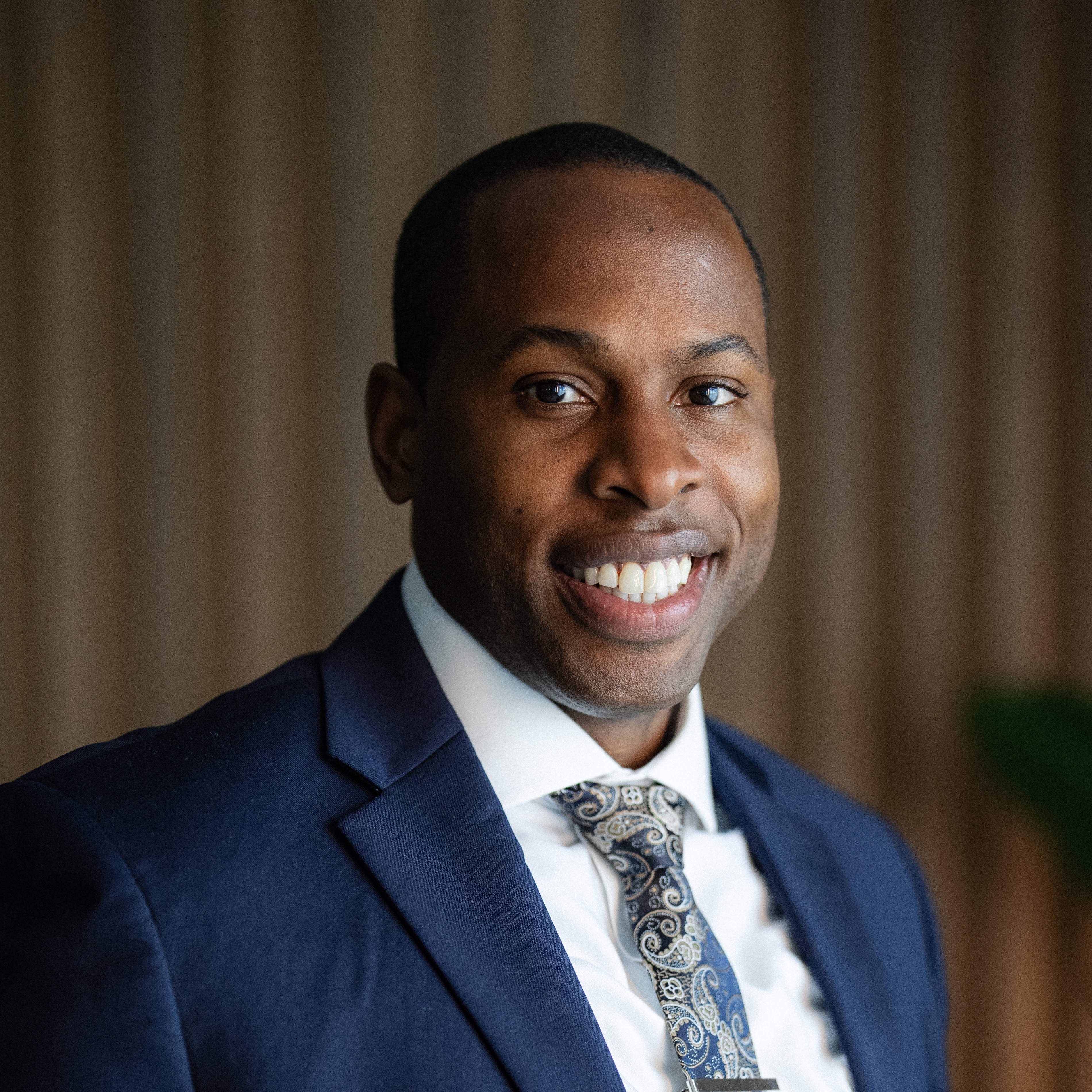 Smiling Black man wearing a navy blue suit jacket, white shirt, and paisley tie against a neutral background.