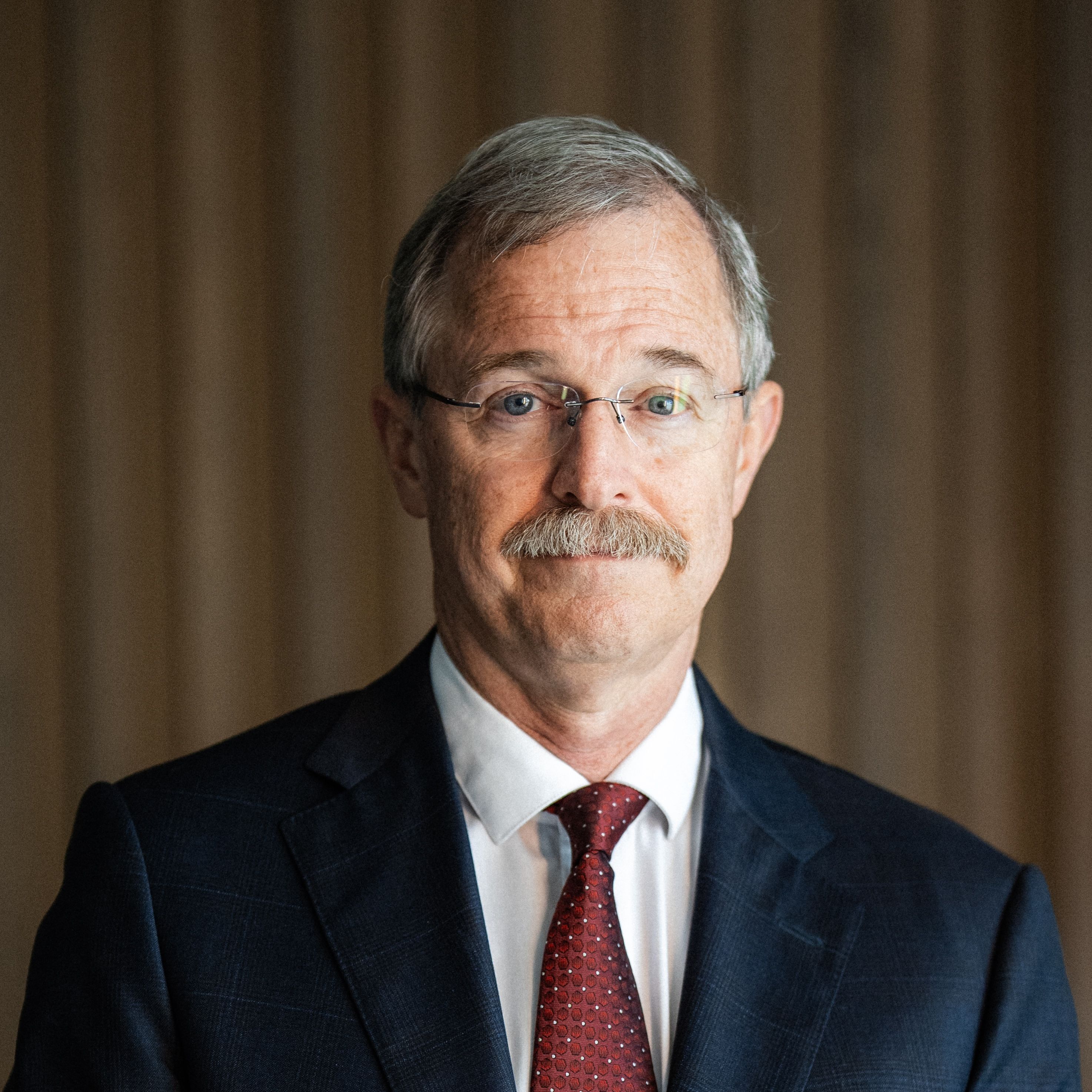 Man with glasses and a mustache wearing a dark suit and red patterned tie against a neutral brown background.