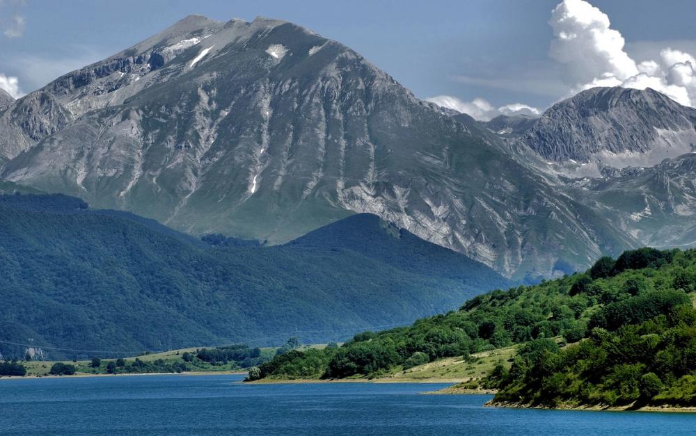 Klart blått vatten framför frodig grön natur med buskar och träd nära strandkanten. I bakgrunden syns flera berg, några låga med skog, och två höga bergstoppar som når upp till molnen.