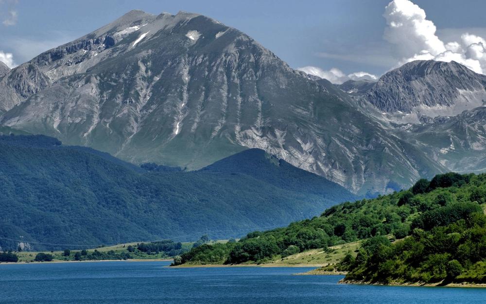 Klart blått vann avbildet foran frodig grønn natur med busker og trær nært vannet. I bakgrunnen er det flere fjell, noen lave med skog vekst, og to høye fjell som rekker opp til skyene helt bak i bildet.