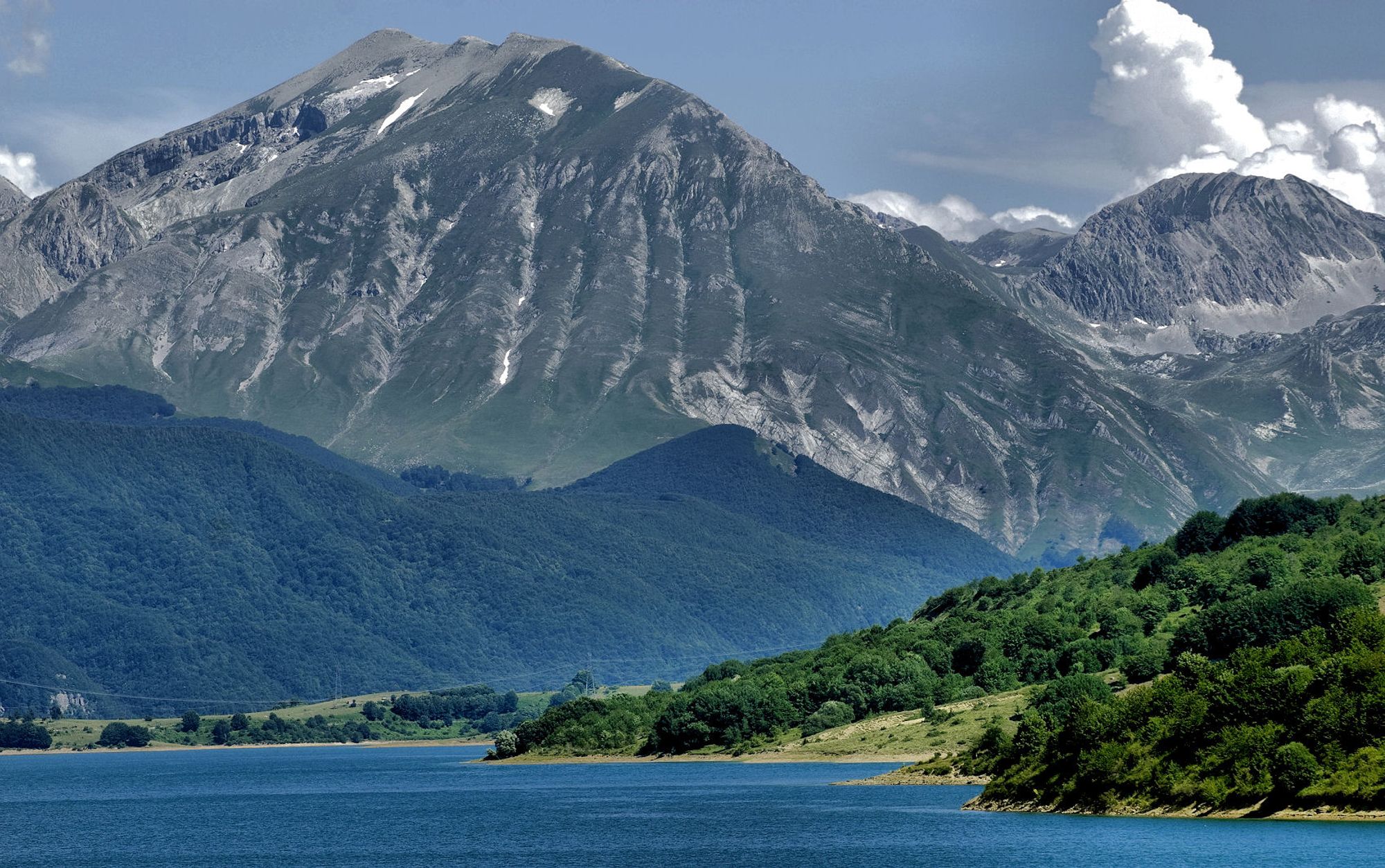 Klart blått vann avbildet foran frodig grønn natur med busker og trær nært vannet. I bakgrunnen er det flere fjell, noen lave med skog vekst, og to høye fjell som rekker opp til skyene helt bak i bildet.