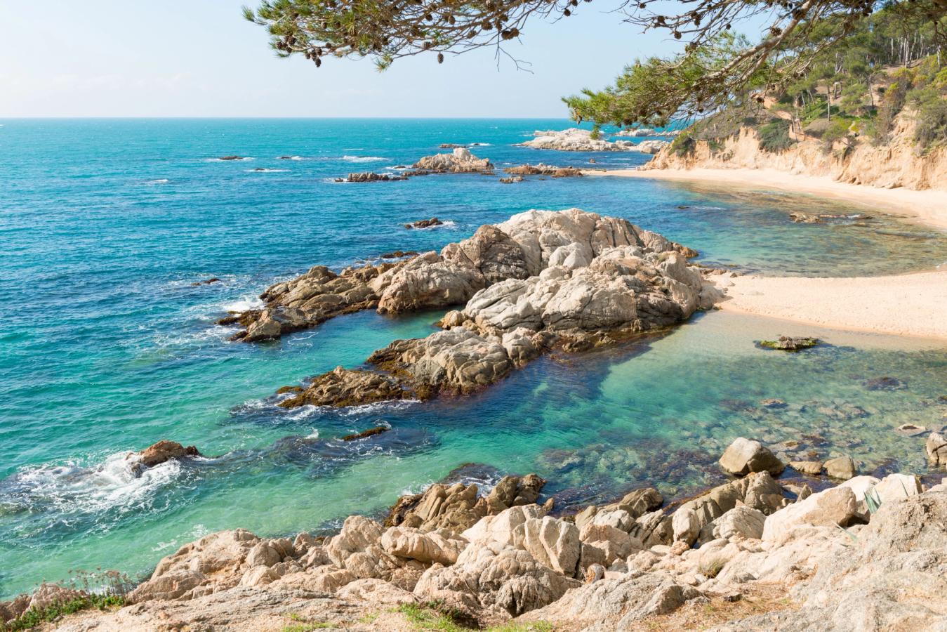 Bildet viser en liten strandbukt på Costa Brava. Finkornet strand på høyre side med små steinformasjoner i vannet.