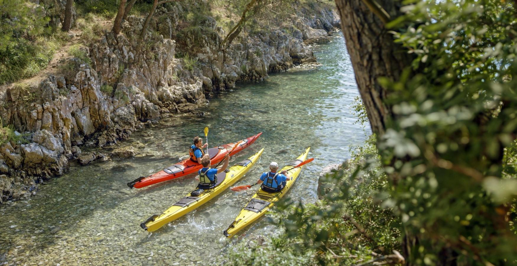 Tre personer i färgglada havskajaker paddlar genom en smal, grund vik omgiven av klippväggar och tallar. Vattnet är klart och grunt med synliga stenar på botten, och naturen runtom är frodig och grön.