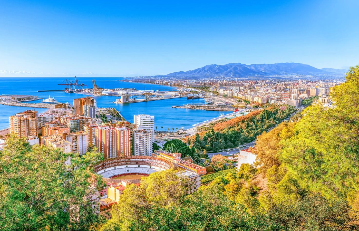 Bildet viser utsikten over Malaga, fra fjellet Gibralfaro. Midt i bildet ser man tyrefekterarenaen Plaza de Toros La Malagueta med den nyere bydelsbebyggelsen. Bakenfor ser man havnen og det blå Middelhavet.