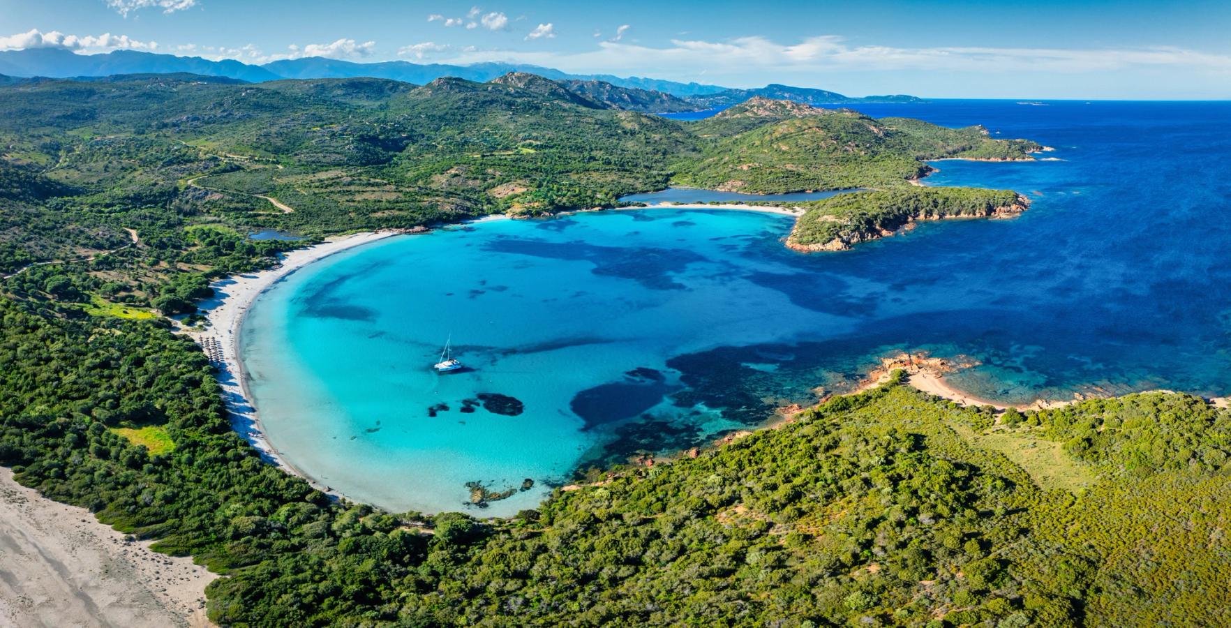 Billedet viser et dronebillede over Rondinara Beach på Korsika. Vi ser en halvcirkel af en bugt med blåt stille vand og en lys strandlinje. Bagved en frodig skov. Himmelen er blå.