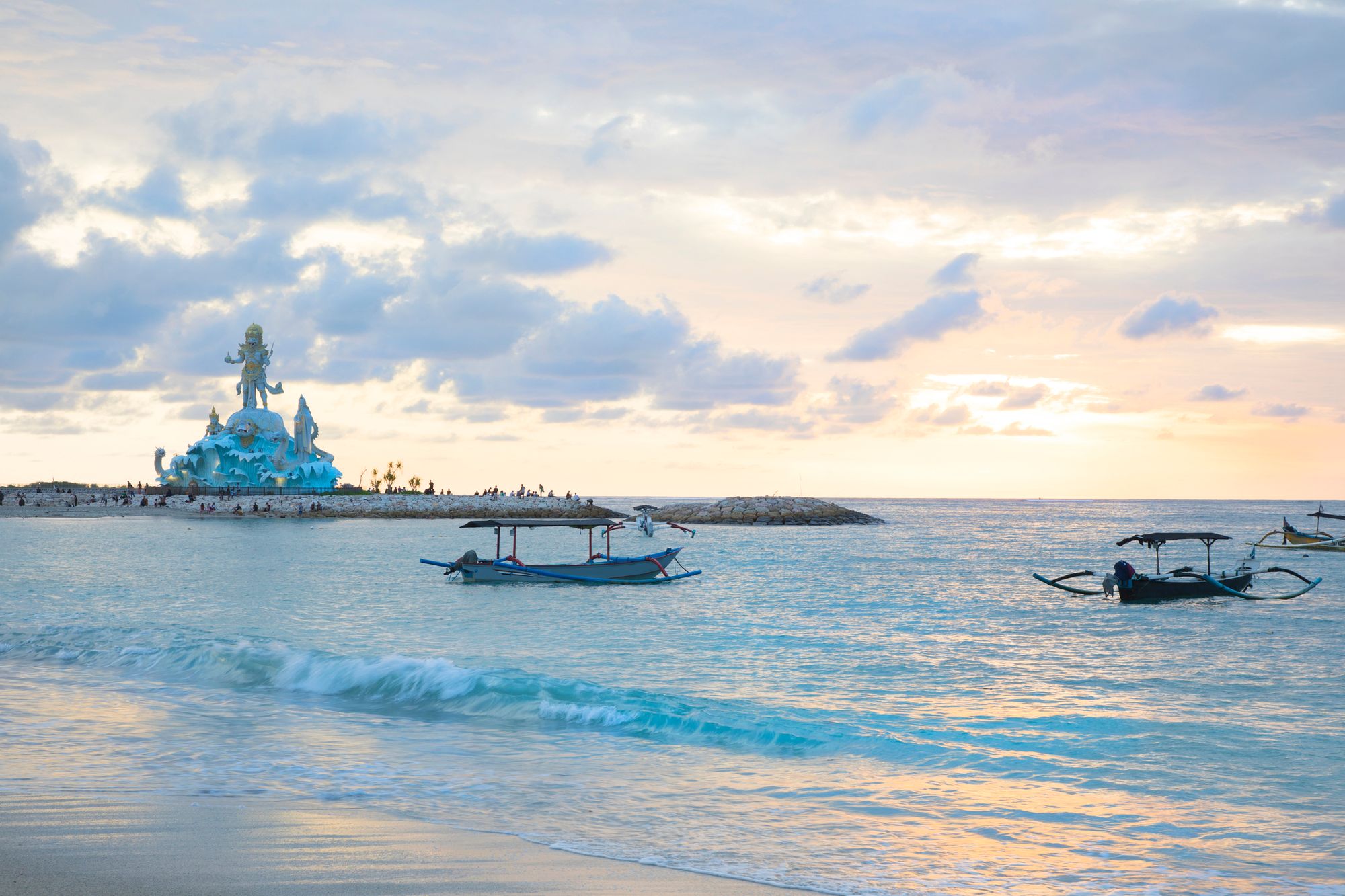 Traditionelle balinesiske fiskebåde i vandet langs Kuta Beach. I baggrunden rejser en stor skulptur sig, mens solen er ved at gå ned i horisonten.