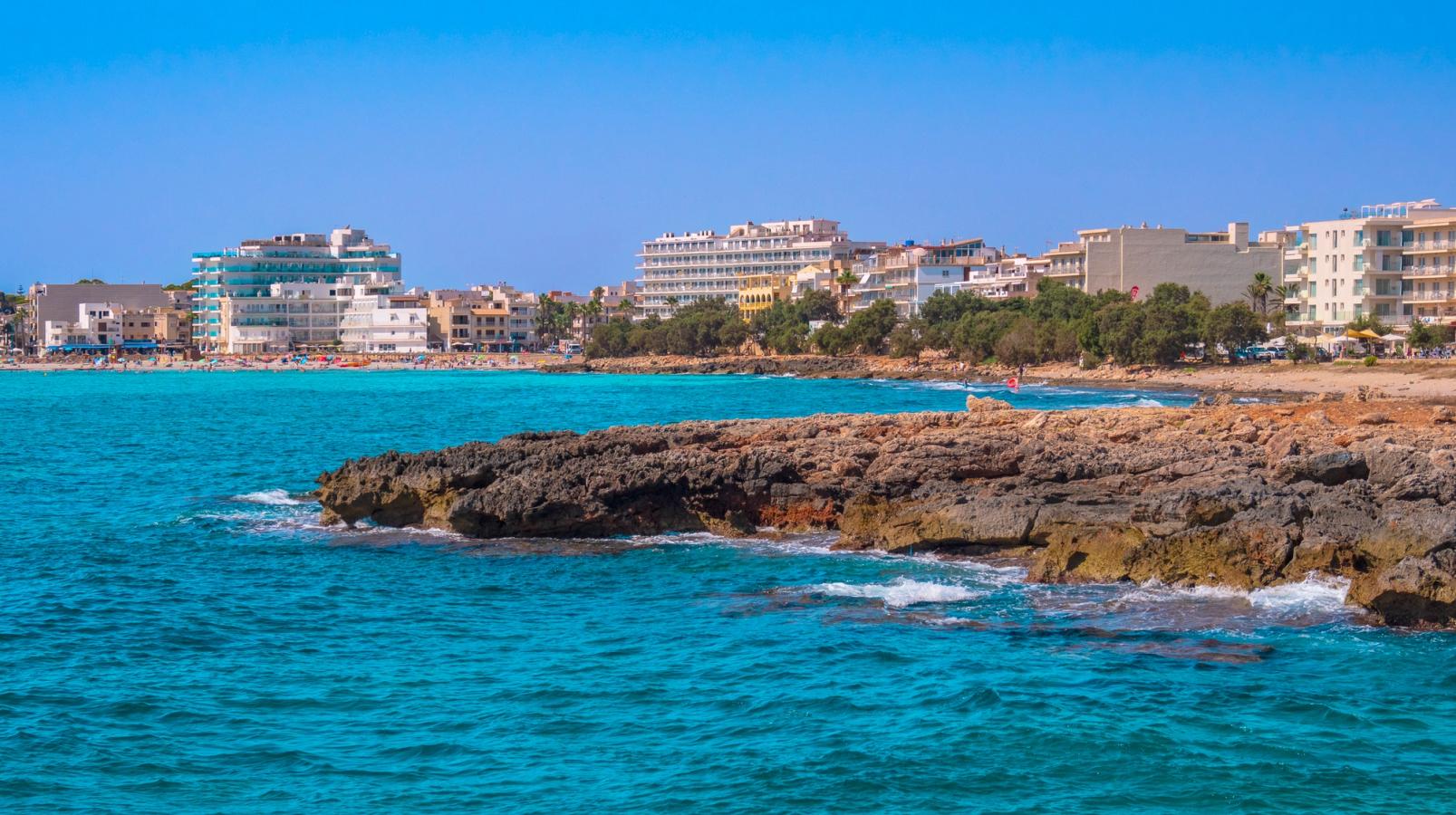  I forgrunden ses en klippe, der rager ud i det azurblå hav. I baggrunden ligger en bred sandstrand, omgivet af hoteller ud mod vandet. 