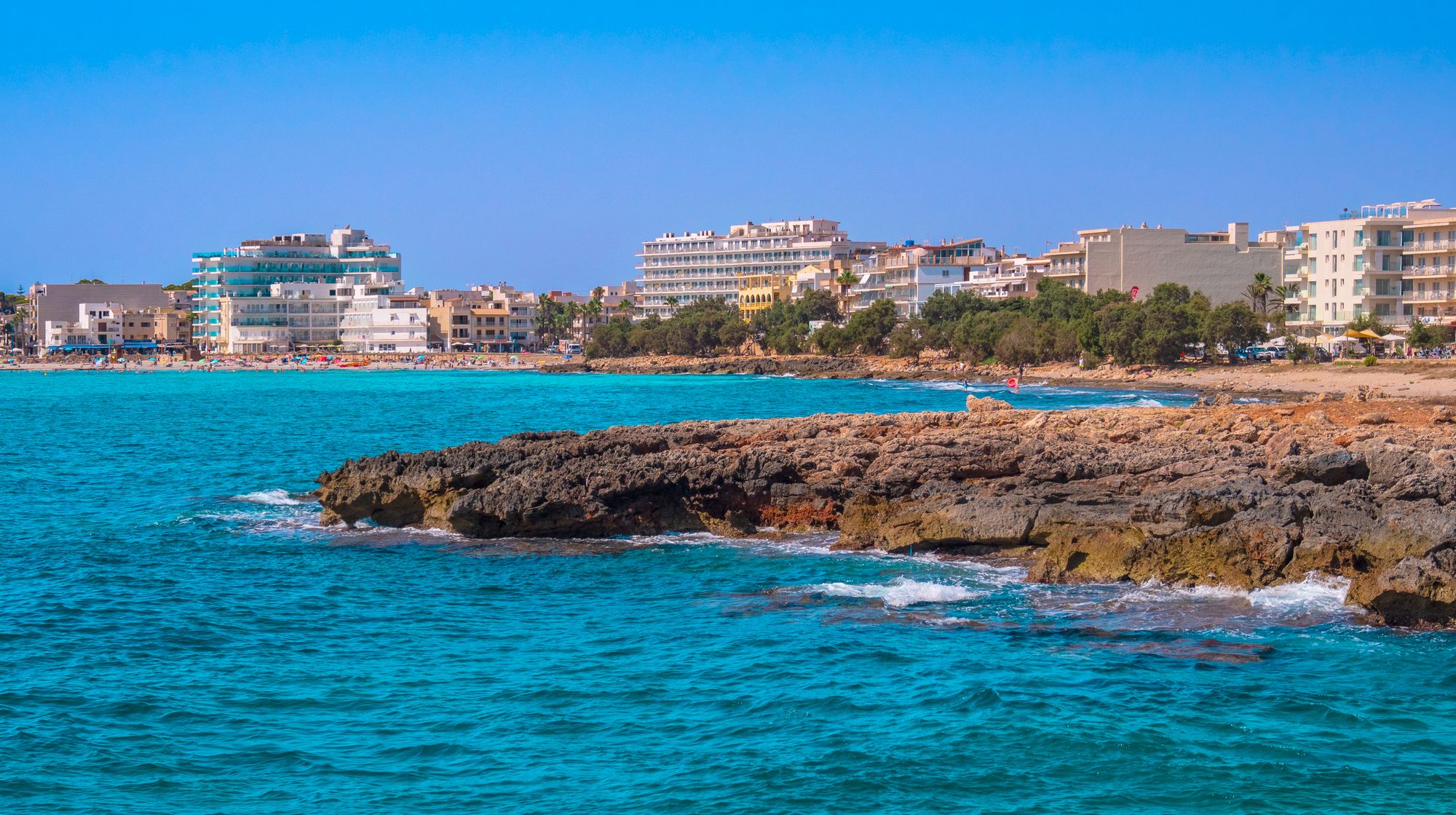 I forgrunden ses en klippe, der rager ud i det azurblå hav. I baggrunden ligger en bred sandstrand, omgivet af hoteller ud mod vandet.