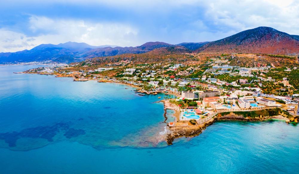Bildet viser byen Hersonissos på Kreta sett ovenifra. Vi ser et blikkstille hav som beveger seg innover mot strandsonen av byen. Byen har spredt bebyggelse med grønne lunger som ender bakerst i bildet med fjellformasjoner.